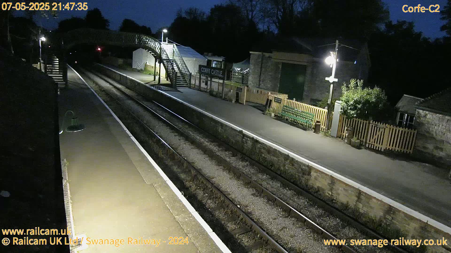 A nighttime view of a deserted train station platform with two sets of tracks, a footbridge, and low lighting.