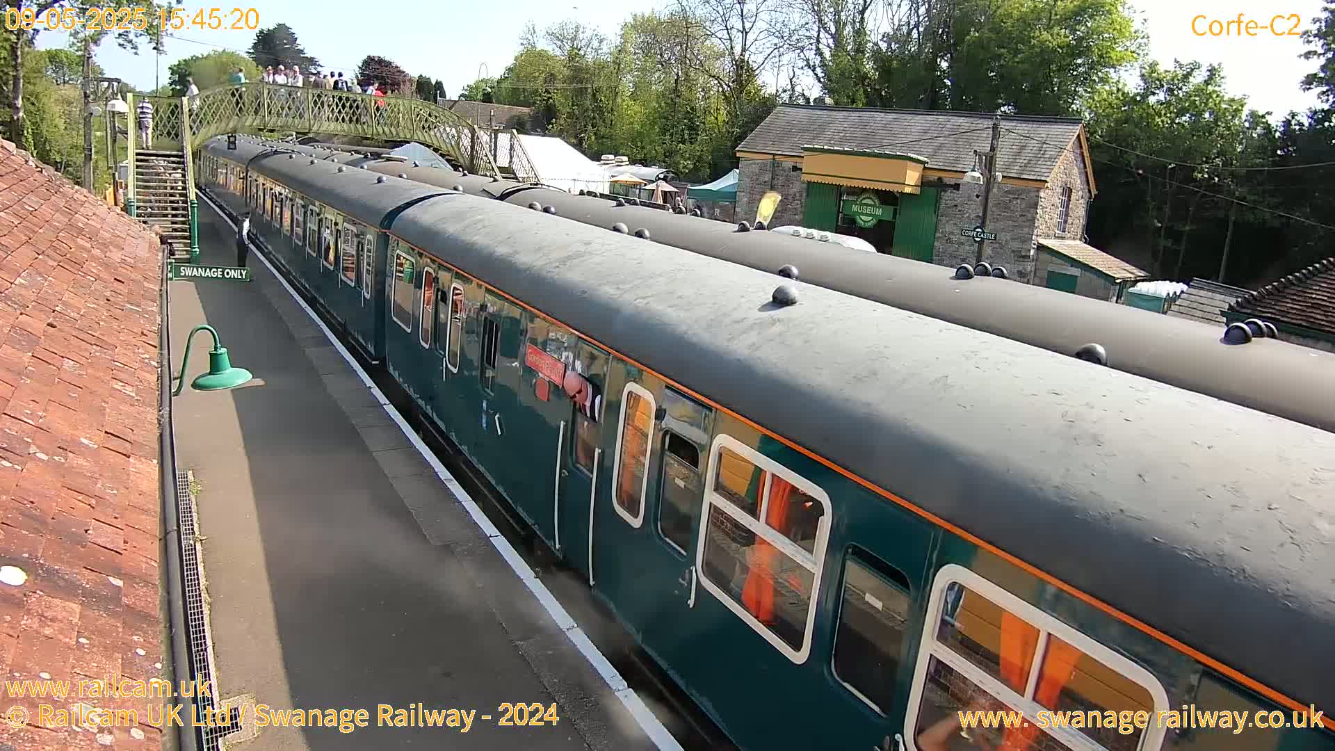 A long, dark-green passenger train sits at a station platform on a sunny day.