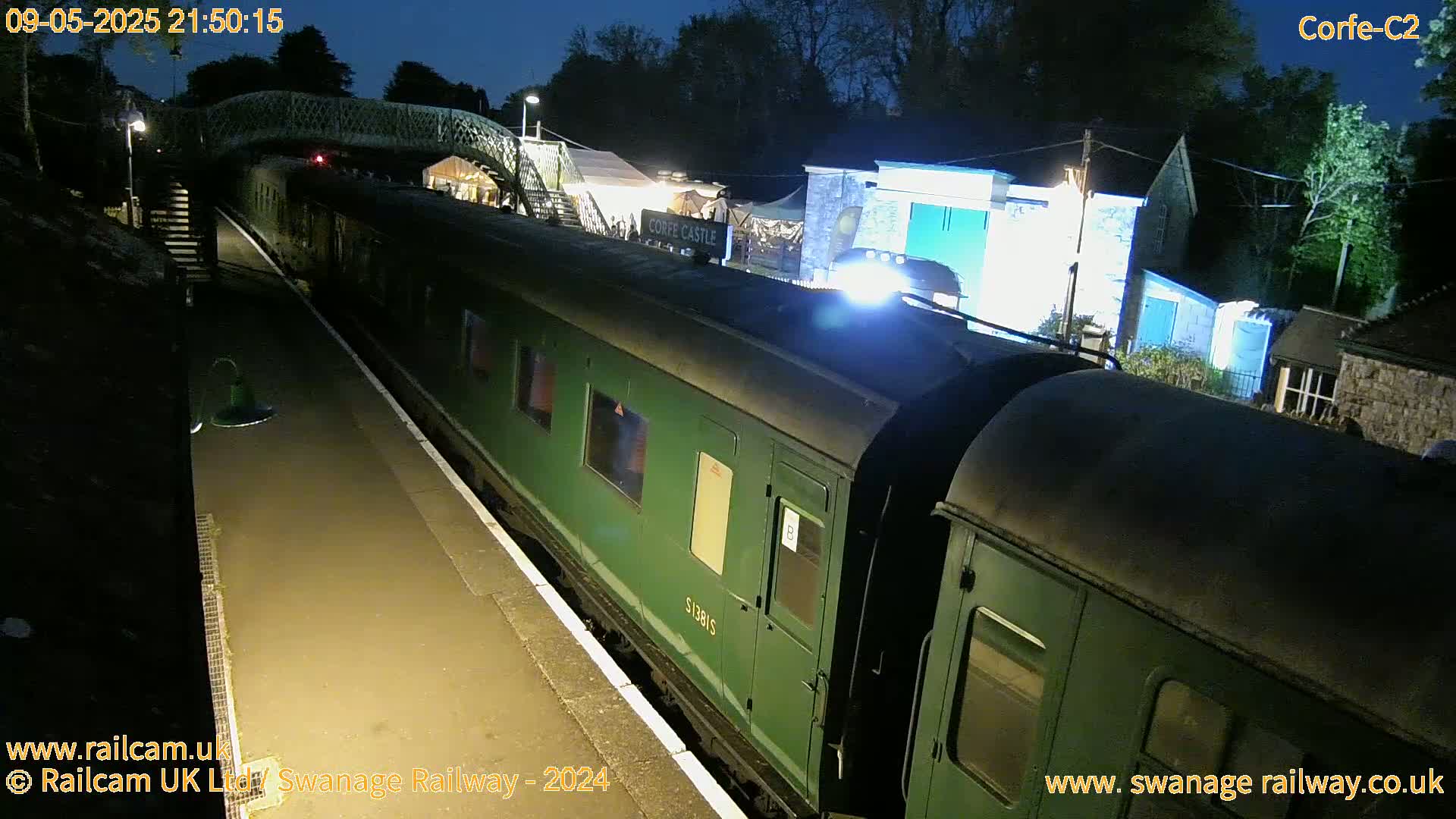 A green passenger train sits at a nighttime station under a footbridge, next to some illuminated buildings.