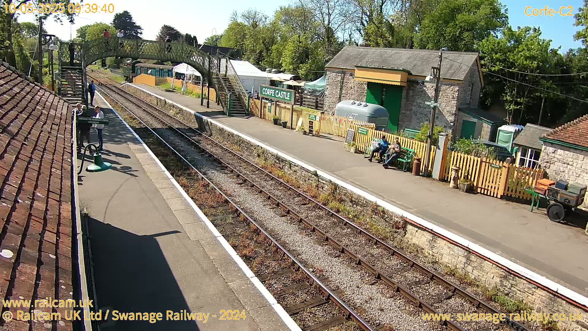 A sunny day at a train station with multiple tracks, a platform, and several buildings, including a small stone structure and a footbridge.