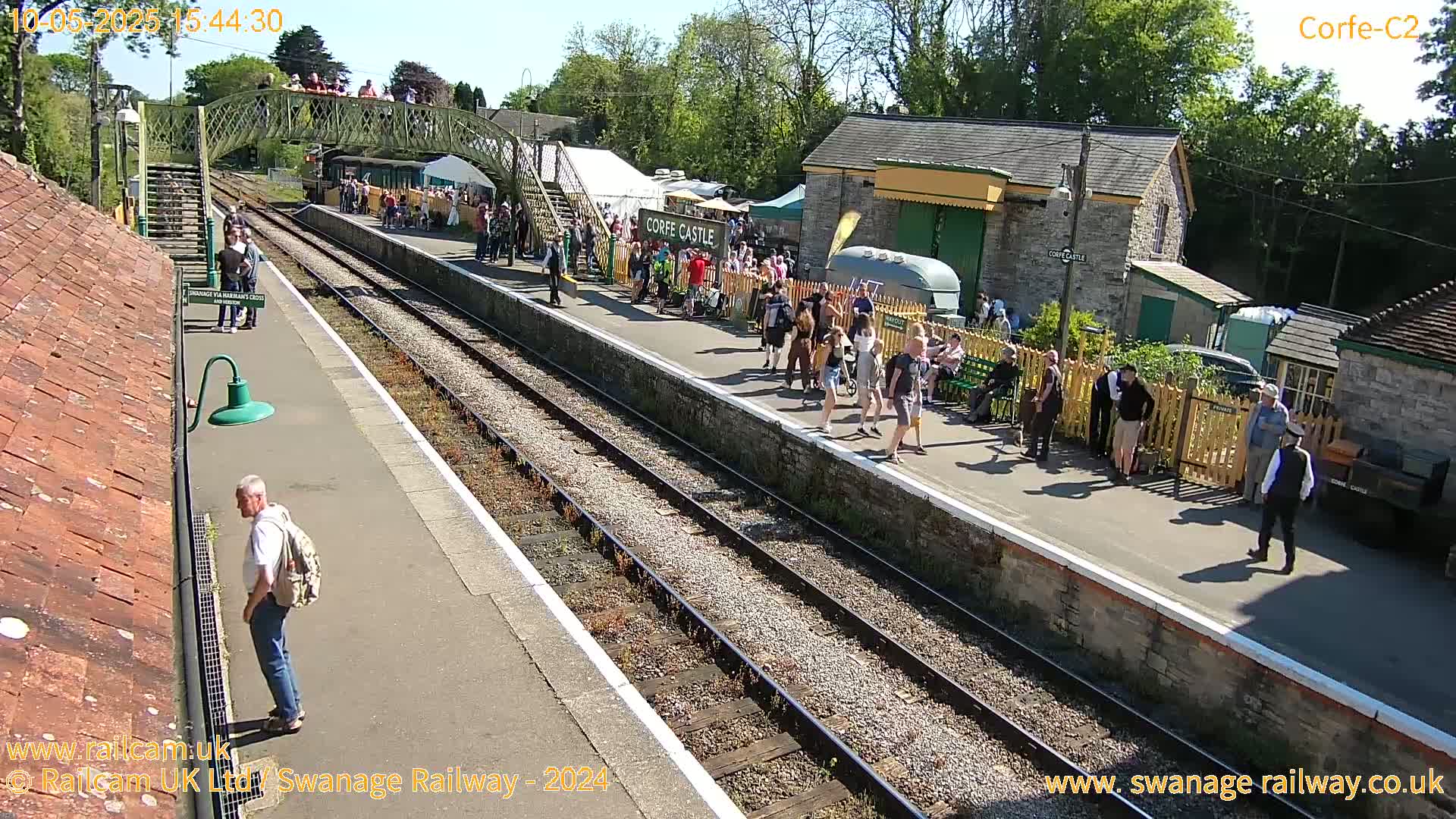 A sunny day at a train station shows a crowd of people gathered on the platform near some stone buildings, with a footbridge overhead.