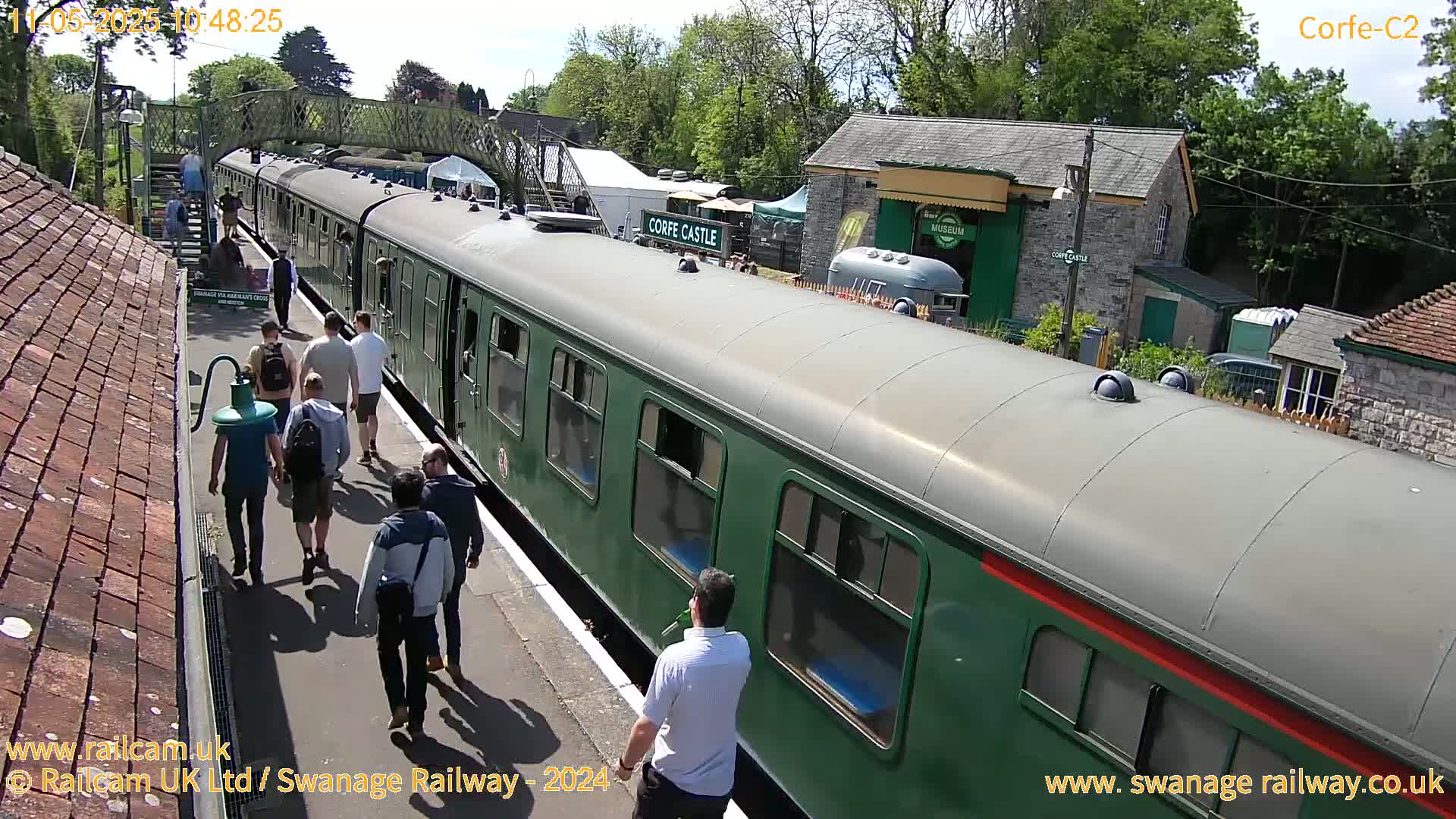 A green passenger train is at a station on a sunny day, with people walking on the platform and buildings visible in the background.
