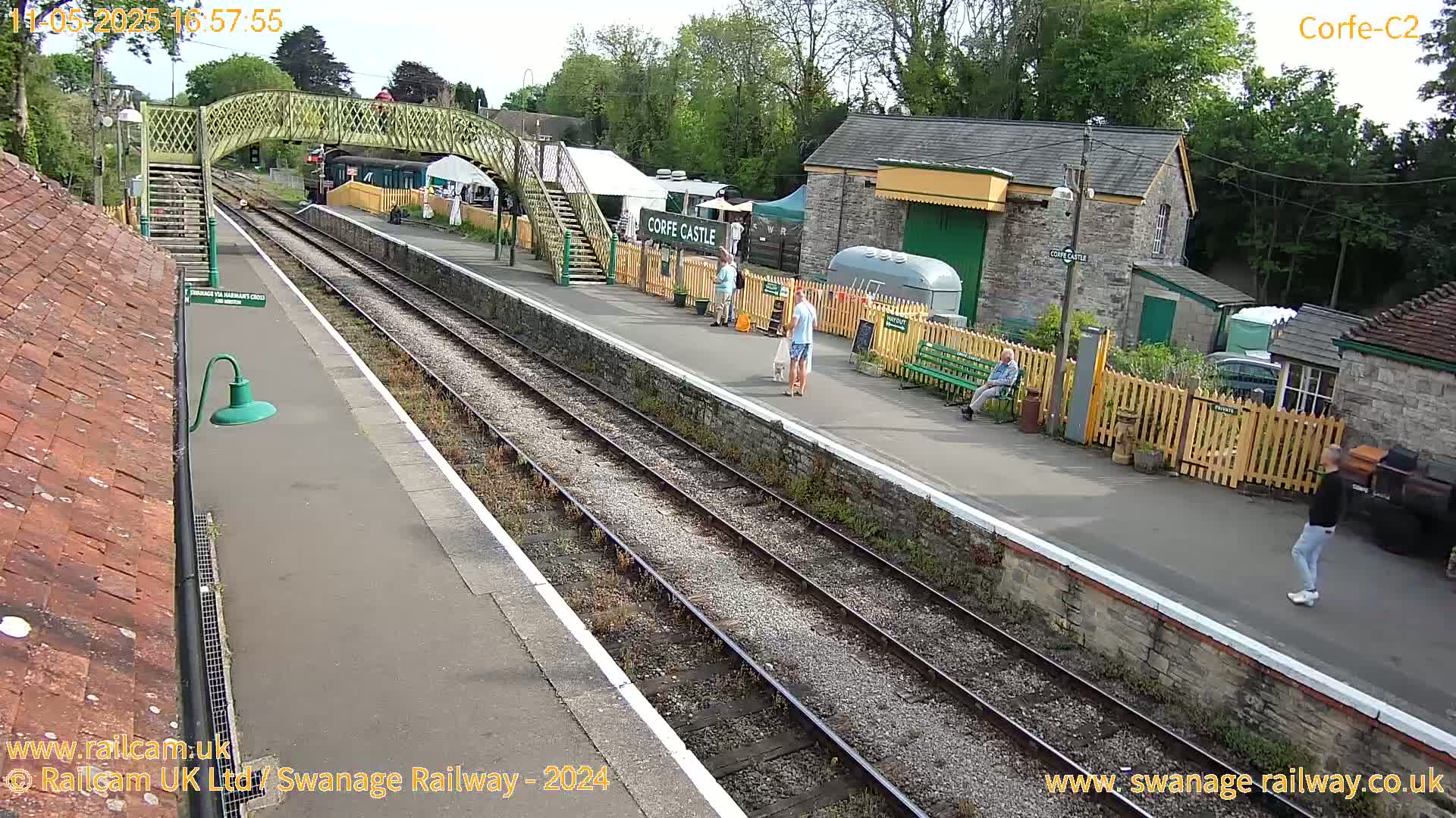 A train station platform with several people, some tents, and railway tracks on a sunny day.