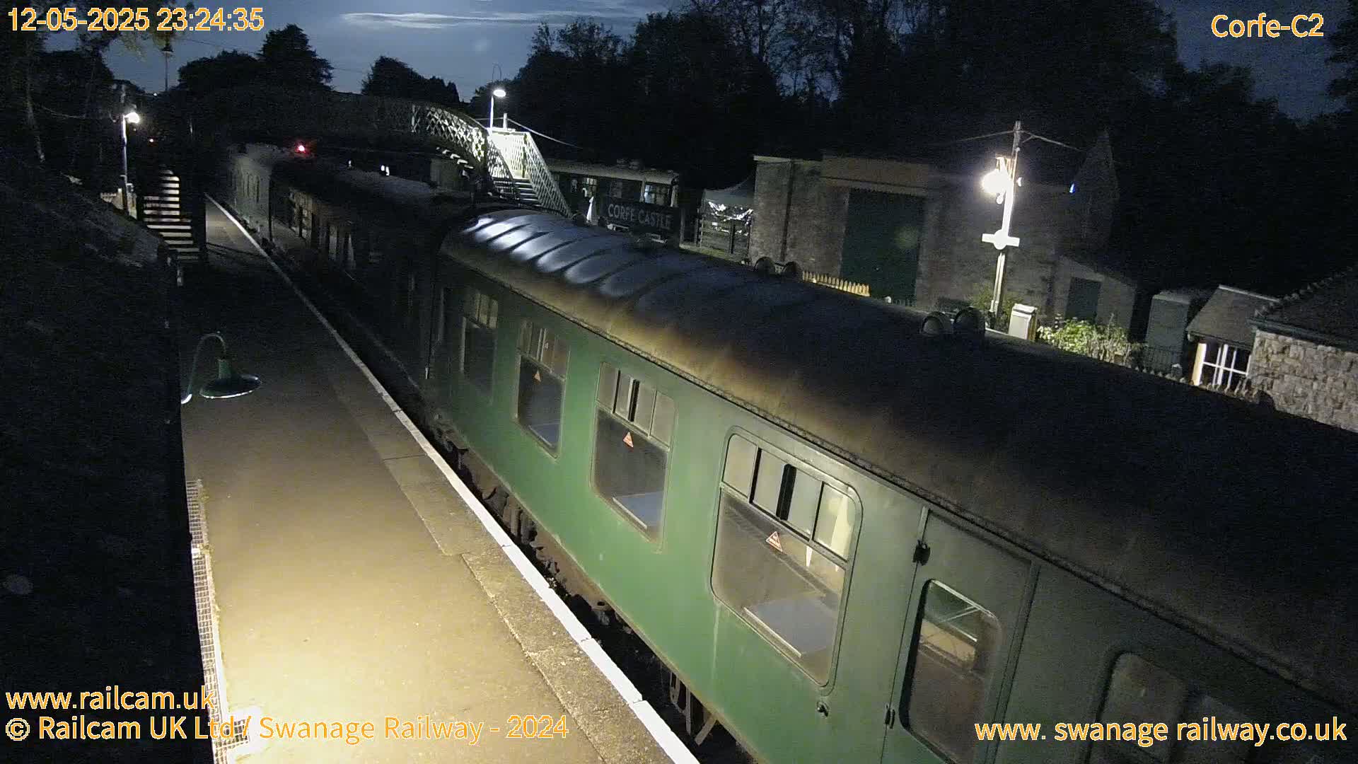 A dark green passenger train sits at a dimly lit nighttime train station under a mostly clear night sky.