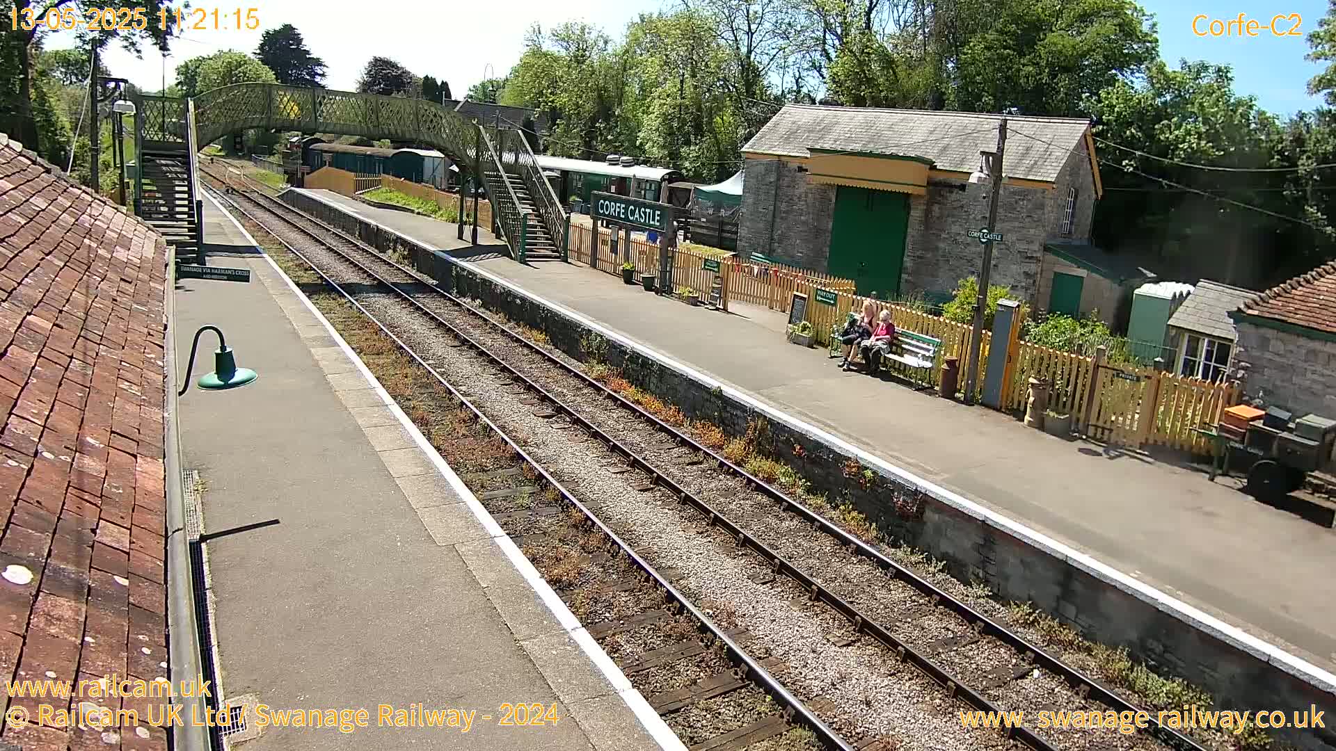 A sunny view of a train station platform with railway tracks, a pedestrian bridge, and several people sitting on a bench.