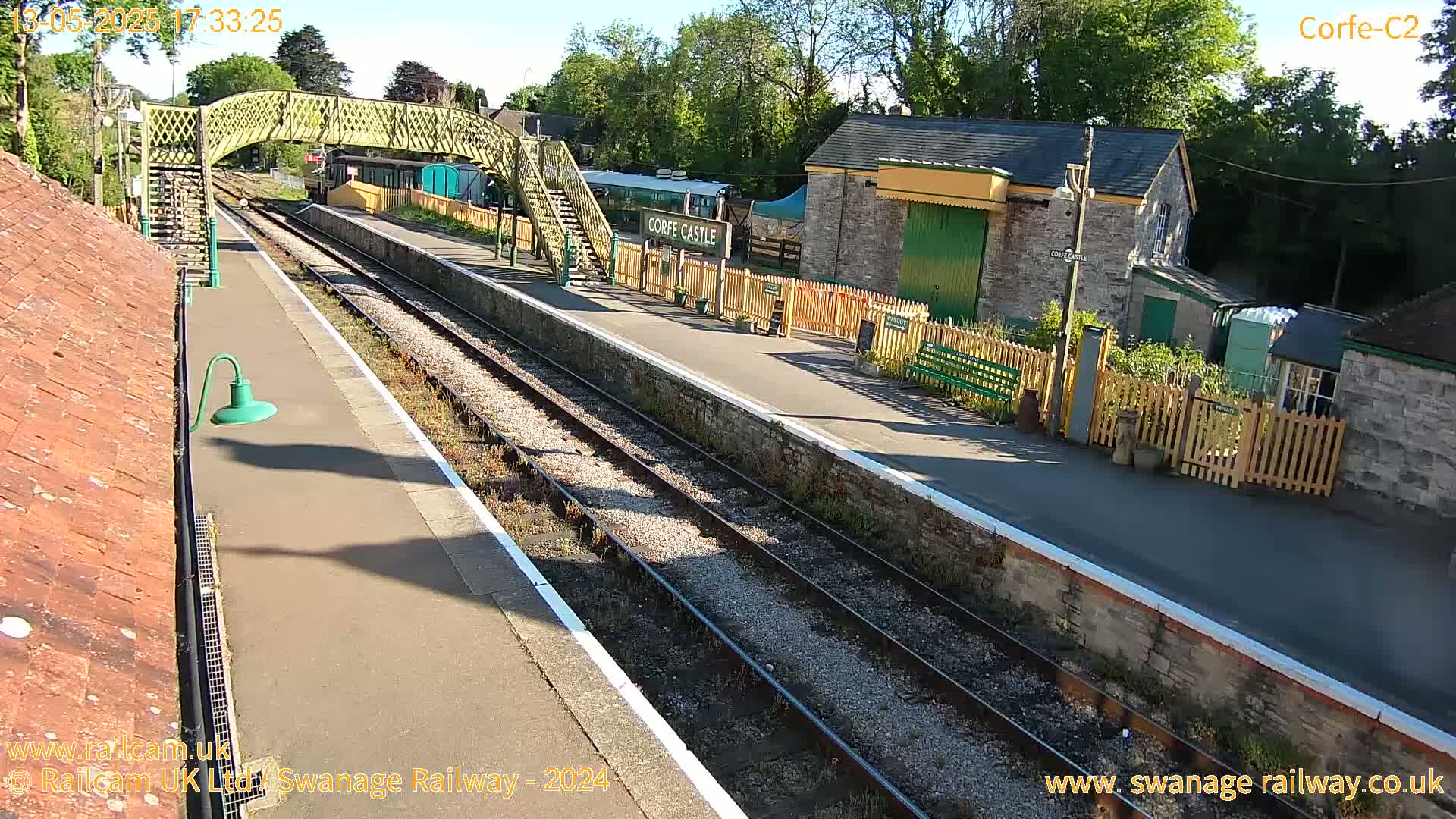 A sunny view of a train station platform with train tracks, a pedestrian bridge, and stone buildings.