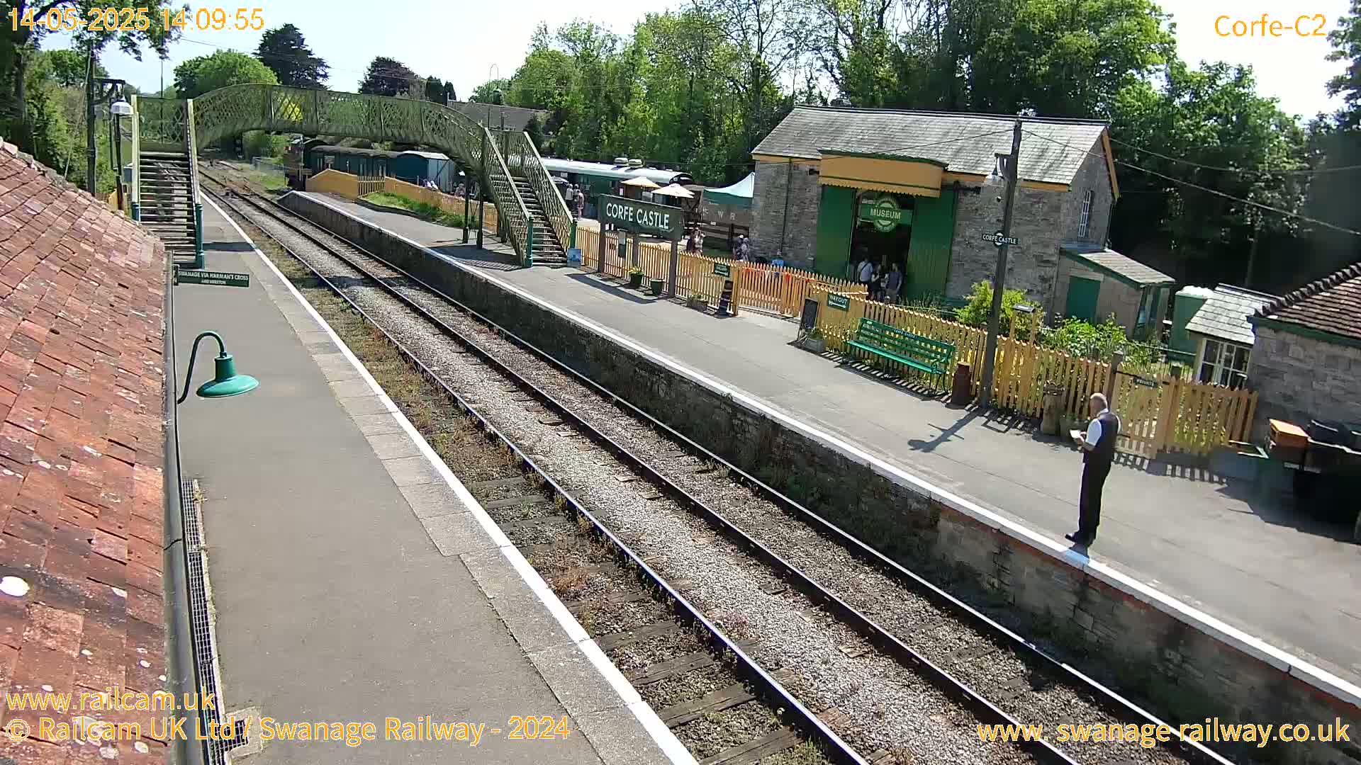 A sunny view of a train station platform with tracks, a small building, and a pedestrian bridge, with several people present.