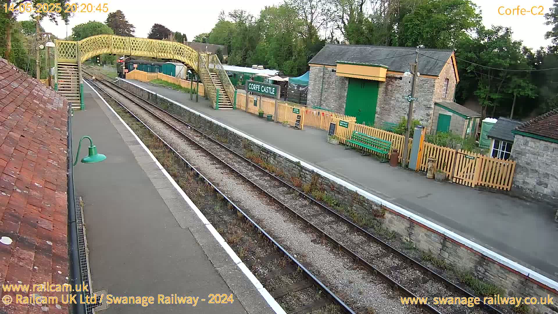 A railway station platform with a yellow footbridge, stone buildings, and train tracks under an overcast sky.