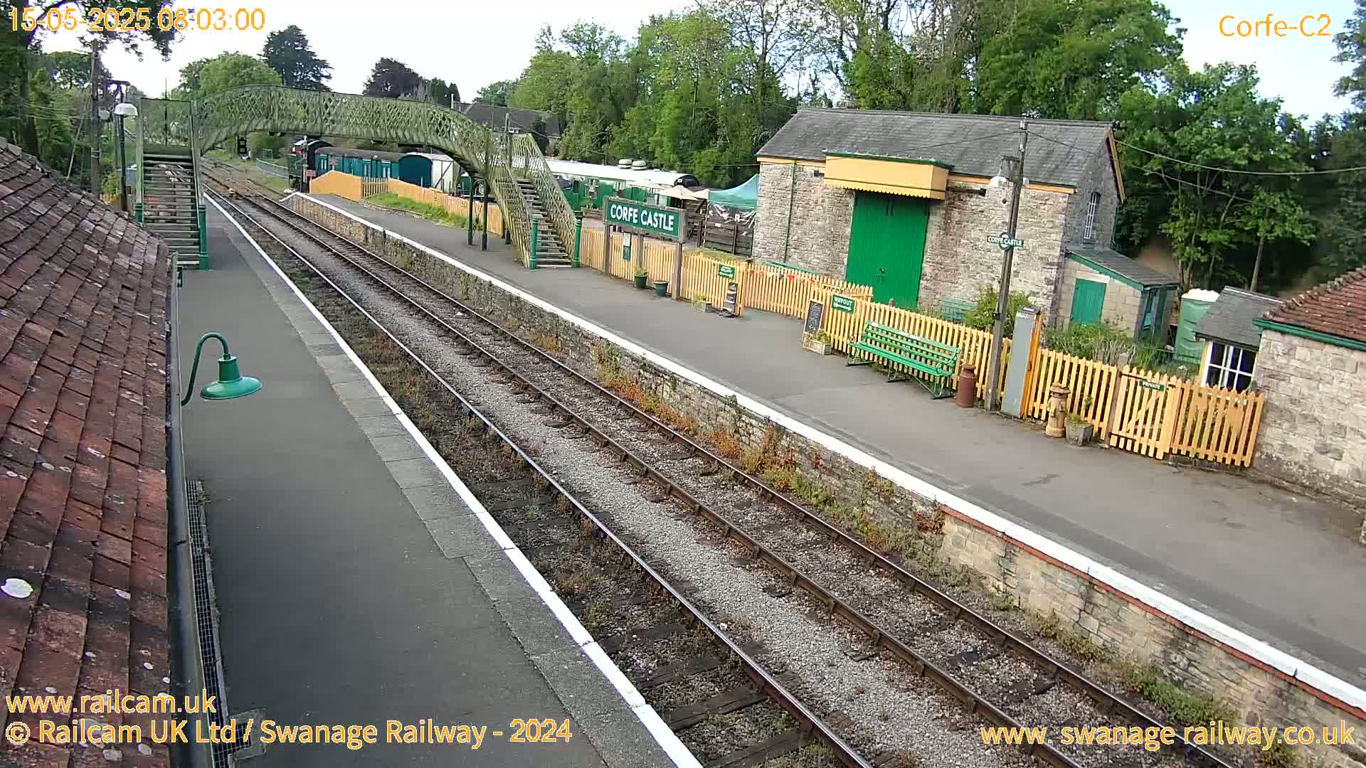 An overcast day at a train station with a footbridge, railway tracks, and stone buildings.