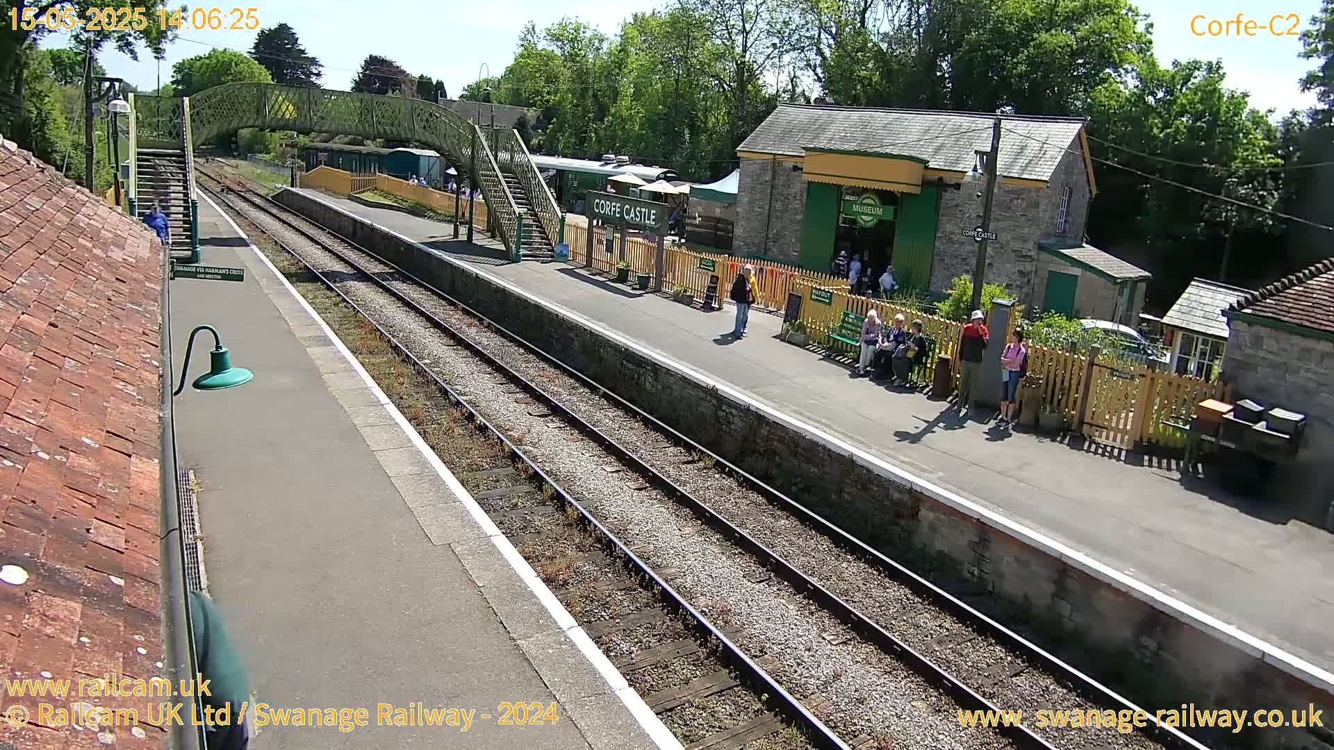 A sunny train station platform with several people standing and sitting near a building,  a train car visible in the background, and train tracks extending into the distance.