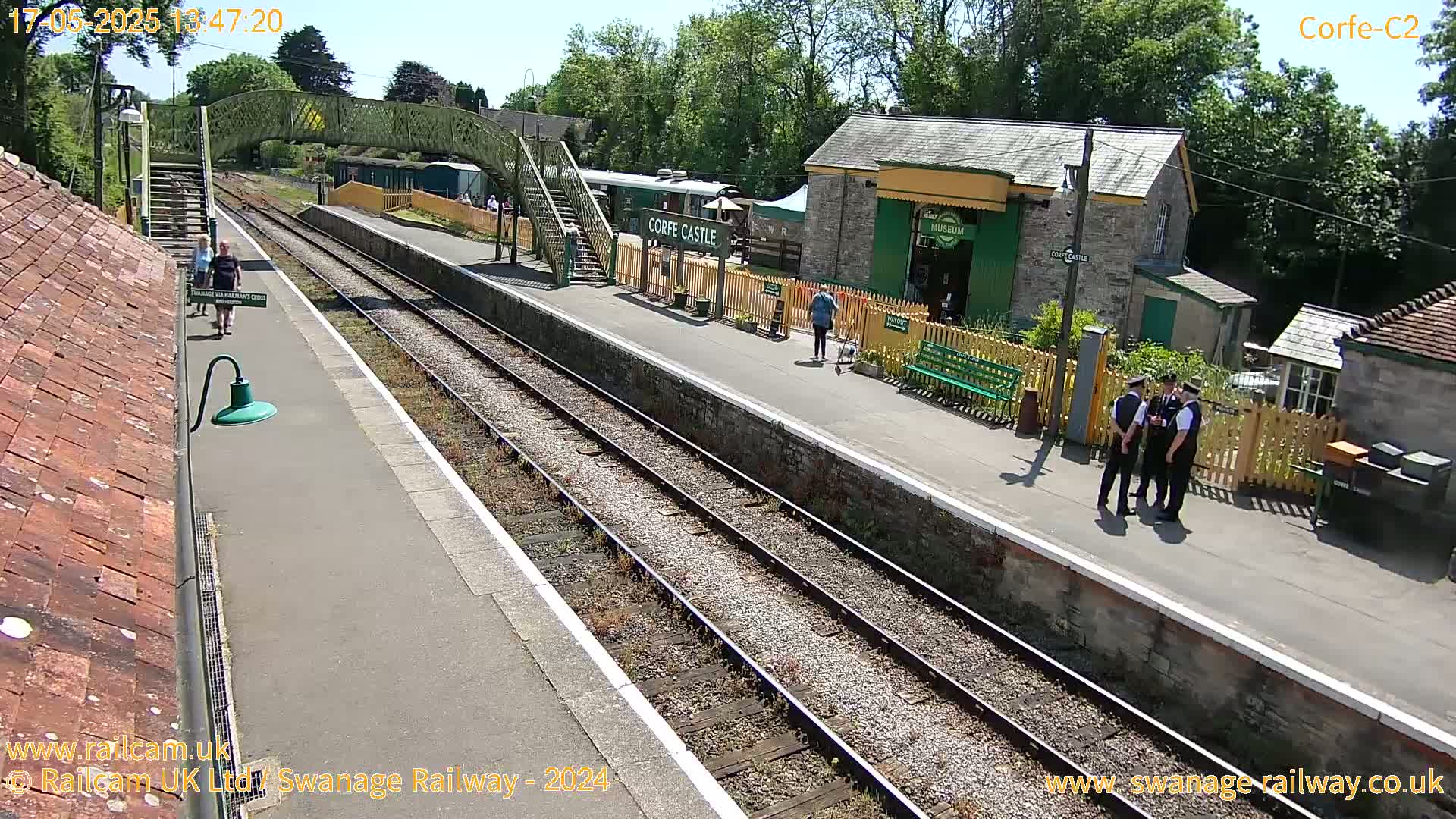 A sunny view of a train station platform with multiple train tracks, a footbridge, and several people.