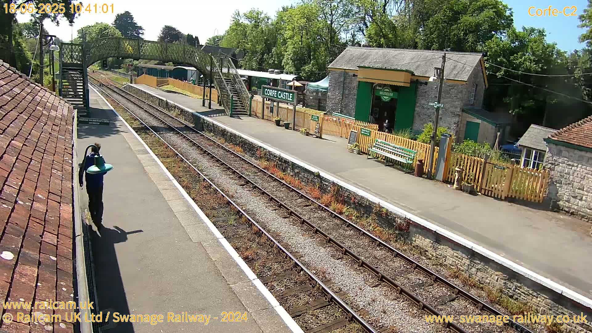 A person carries a green object on a sunny day at a train station with multiple tracks and a small building.