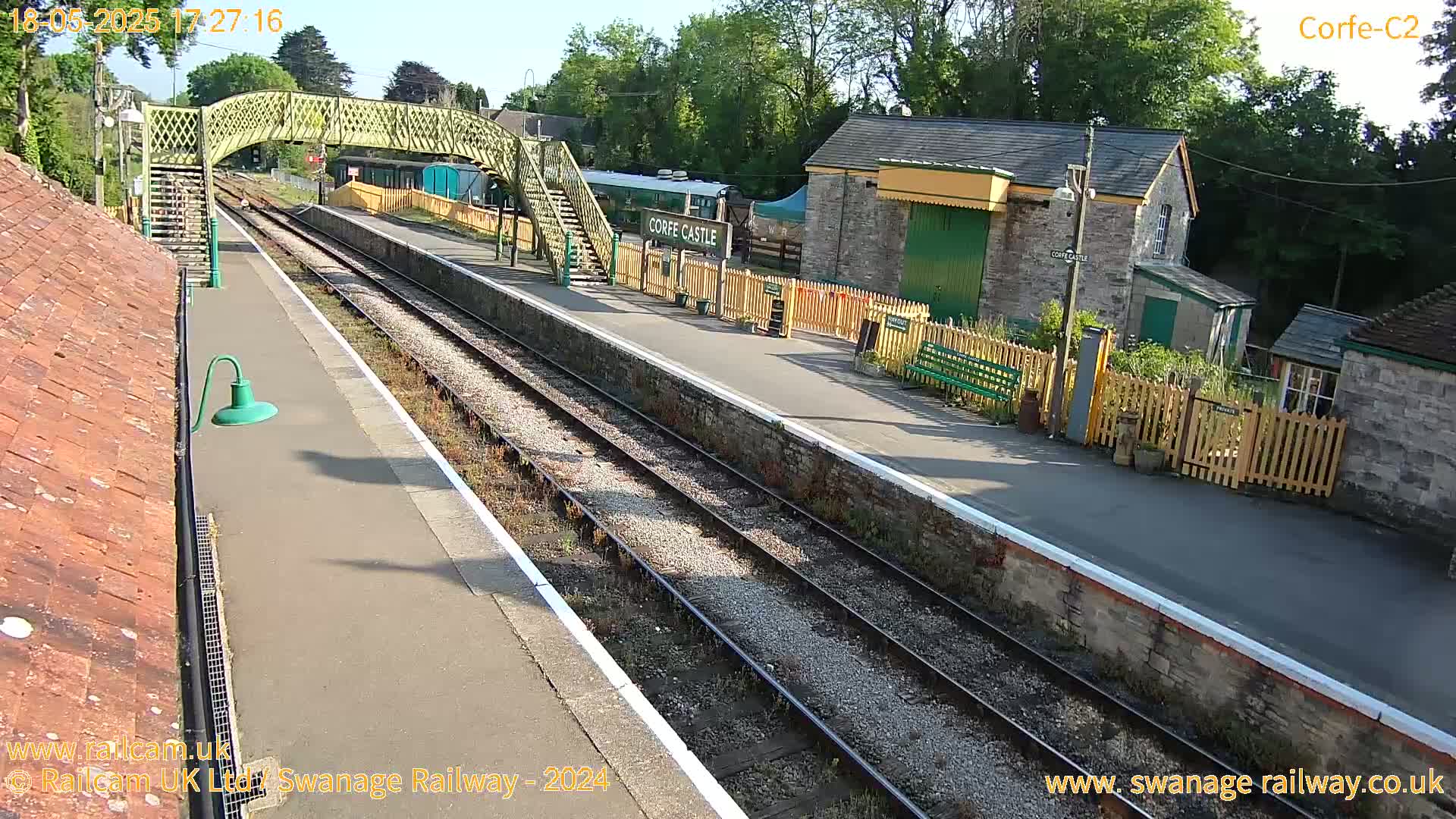 A train station platform with two parallel train tracks, a footbridge, and stone buildings, on a sunny day.