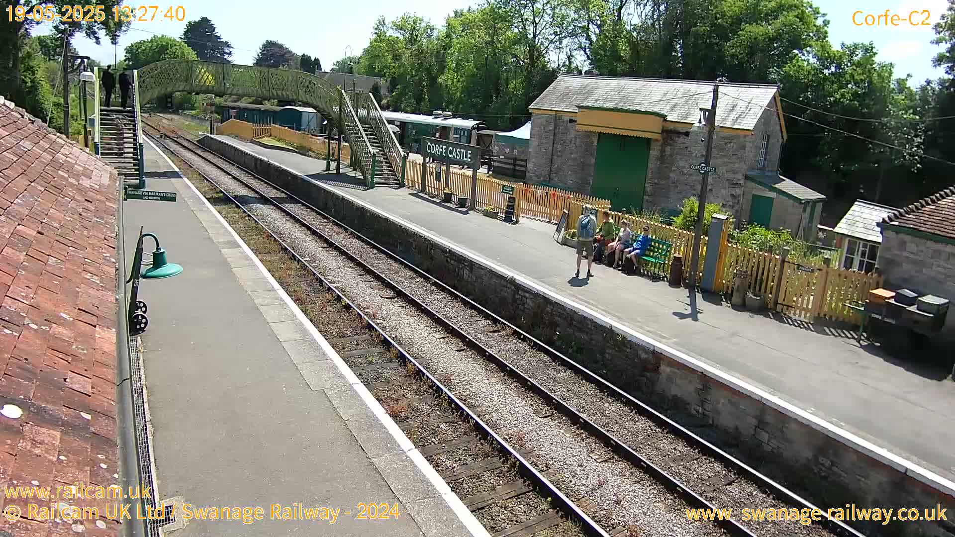 A sunny day at a train station with multiple train tracks, a platform, a footbridge, several buildings, and a few people standing and sitting near a wooden fence.
