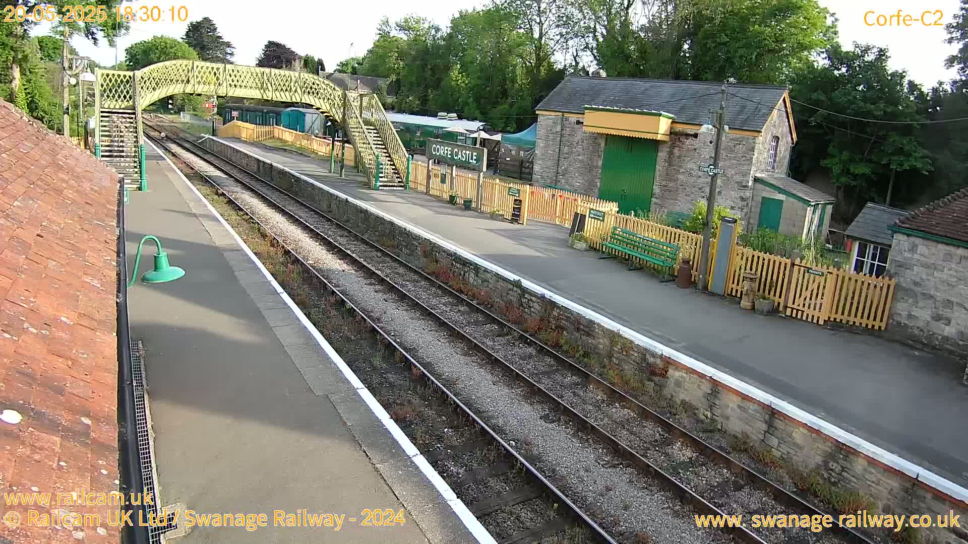 A sunny view of a train station with a footbridge, tracks, and several buildings, including a stone structure.