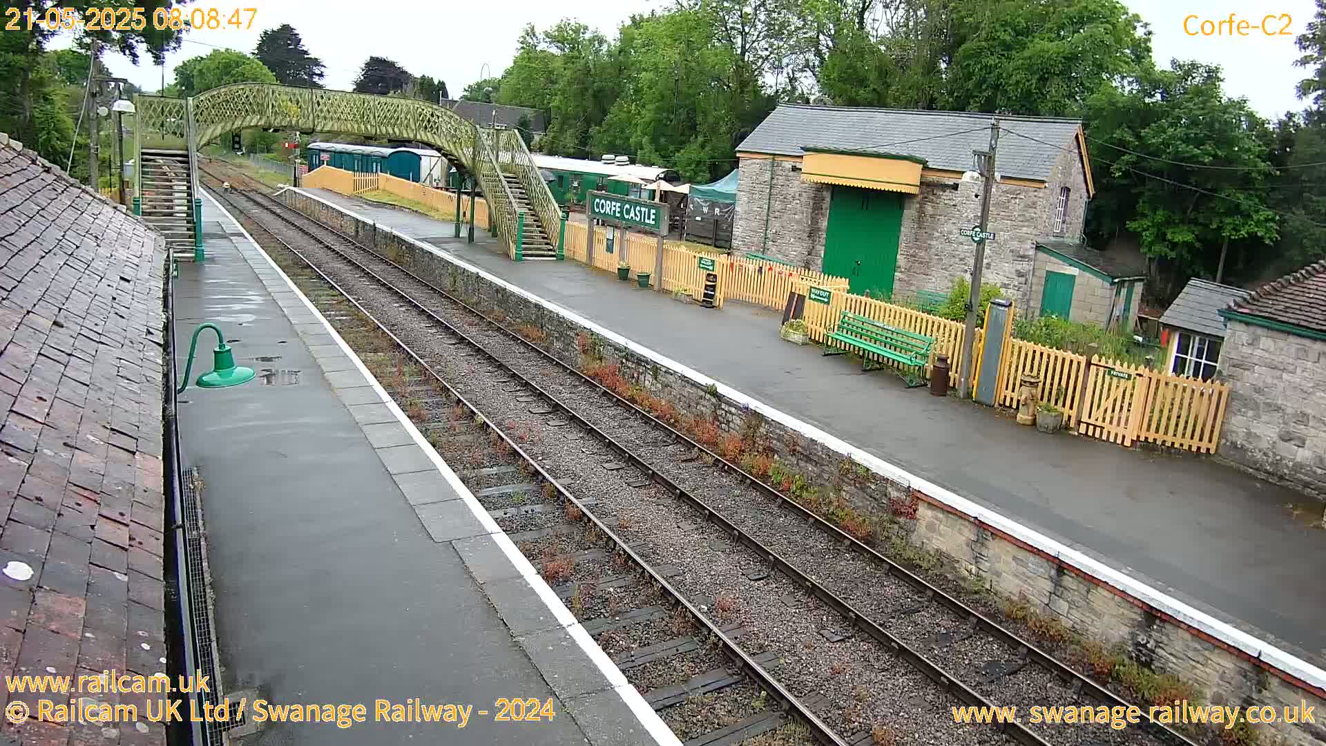 A wet train station platform with a pedestrian bridge and several stone buildings is visible under an overcast sky.