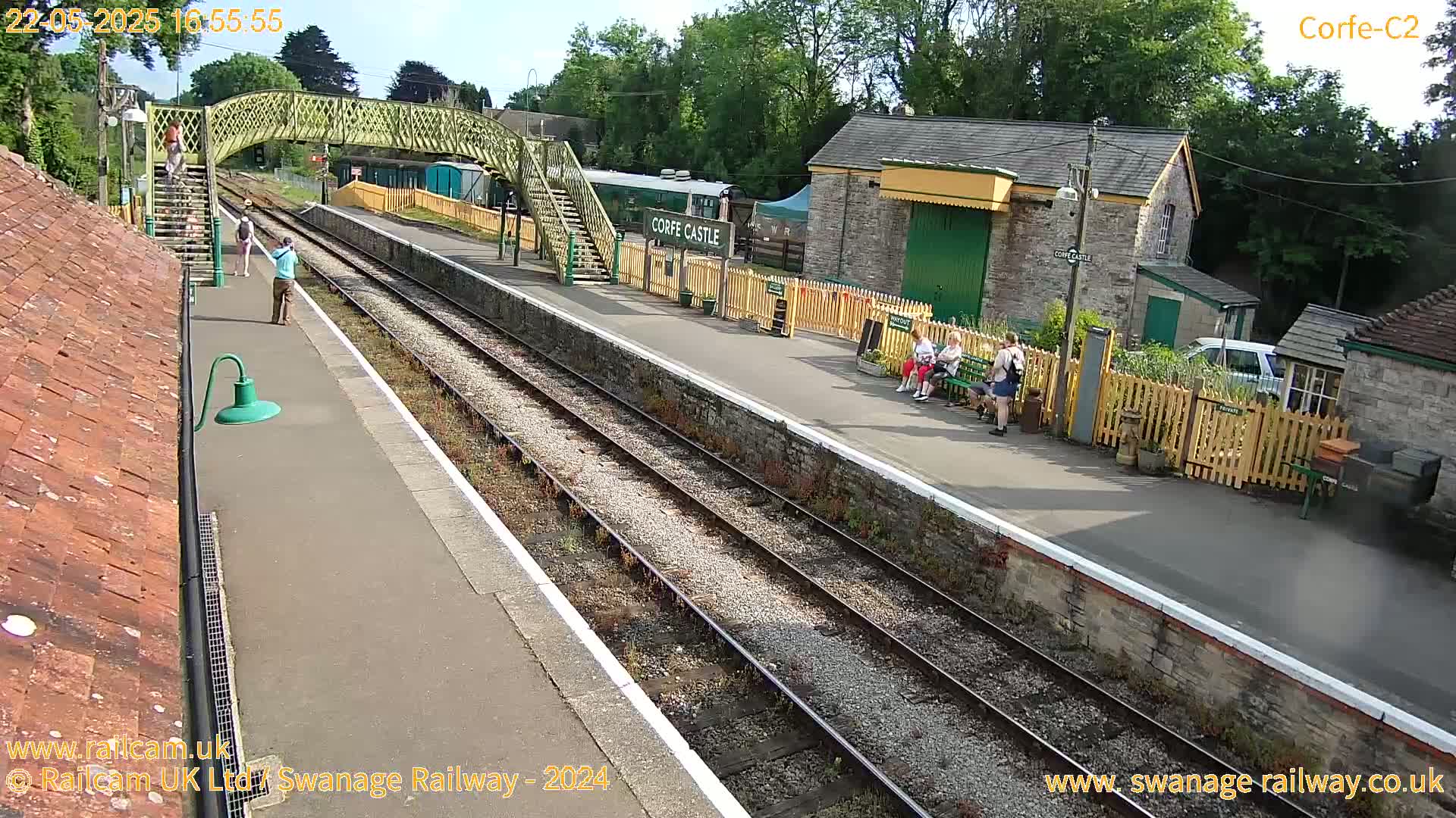 A high-angle view of a train station platform on a sunny day shows several people waiting near a stone building and a railway carriage, with a footbridge overhead.