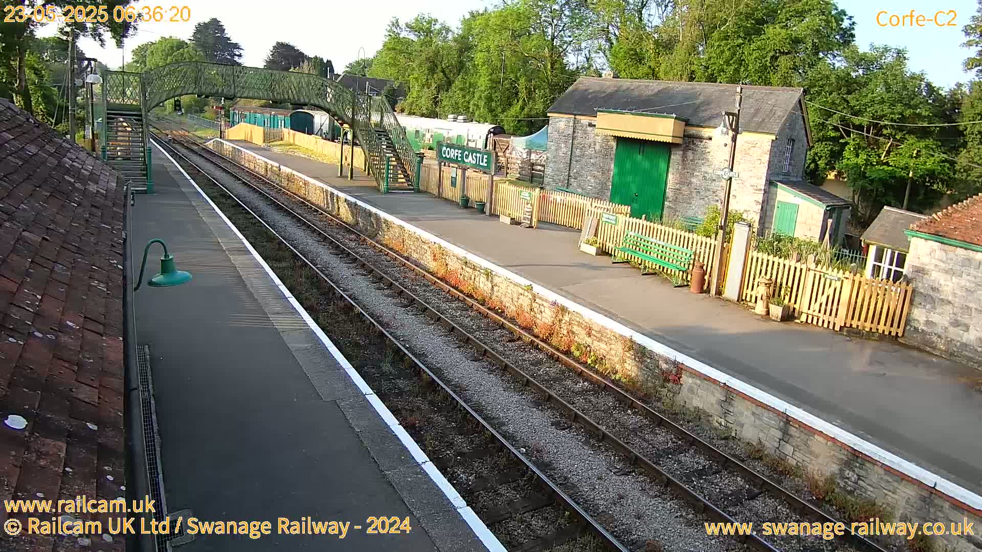 A train station platform with two train tracks, a stone building, and a footbridge under a partly sunny sky.
