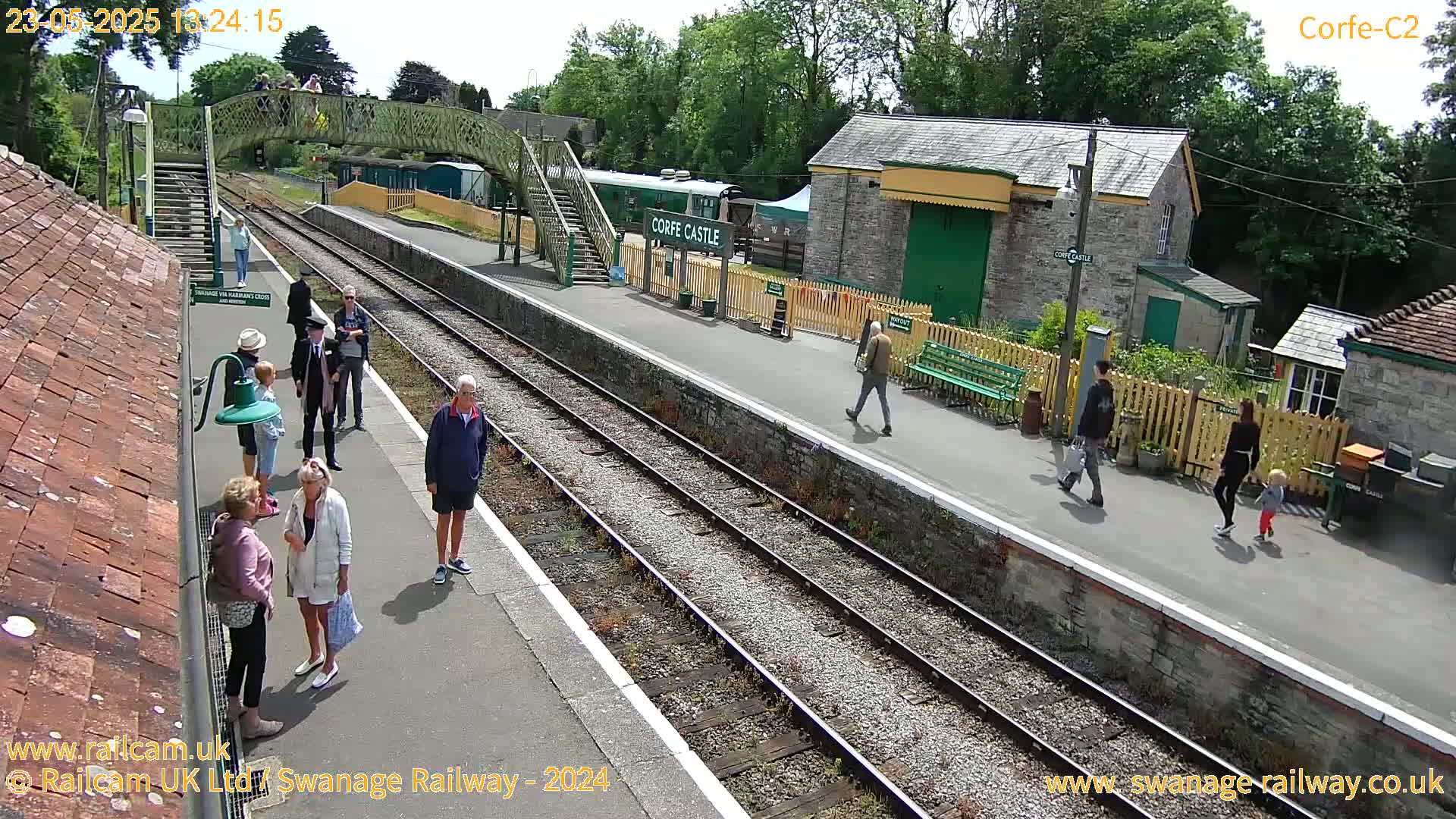 A sunny day at a train station with several people standing on the platform and walking along the tracks, near a green train and a footbridge.