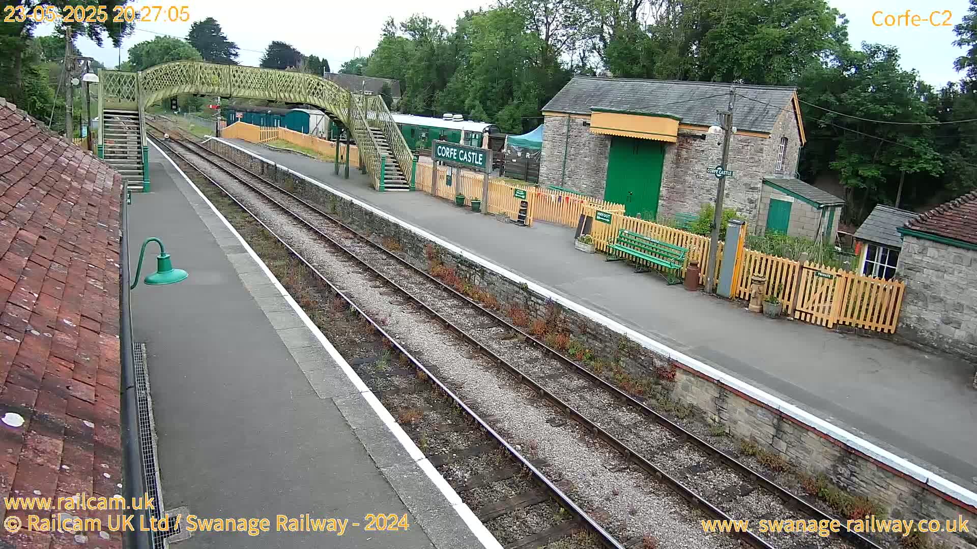 A train station platform with railway tracks, a pedestrian bridge, and several stone buildings is visible on an overcast day.