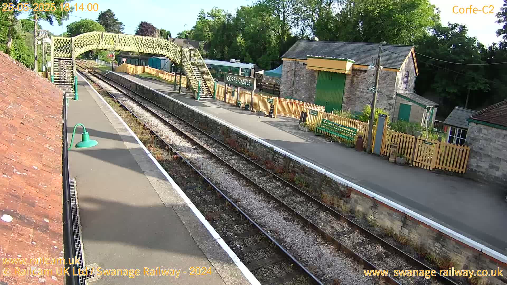 A sunny view of a train station with a footbridge, platform, and stone buildings.