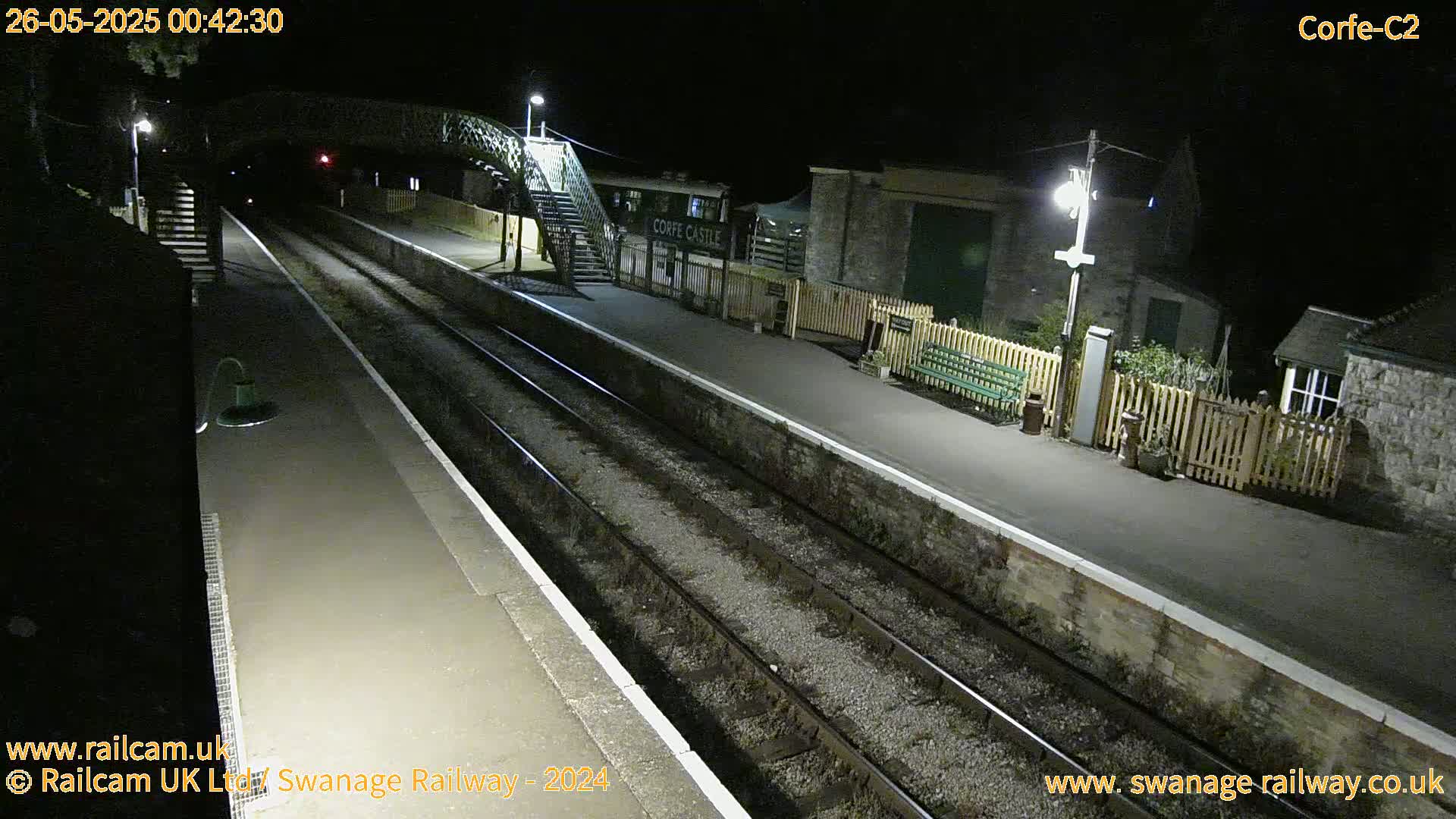 A nighttime view of a deserted train station platform with two train tracks, a footbridge, and several buildings.