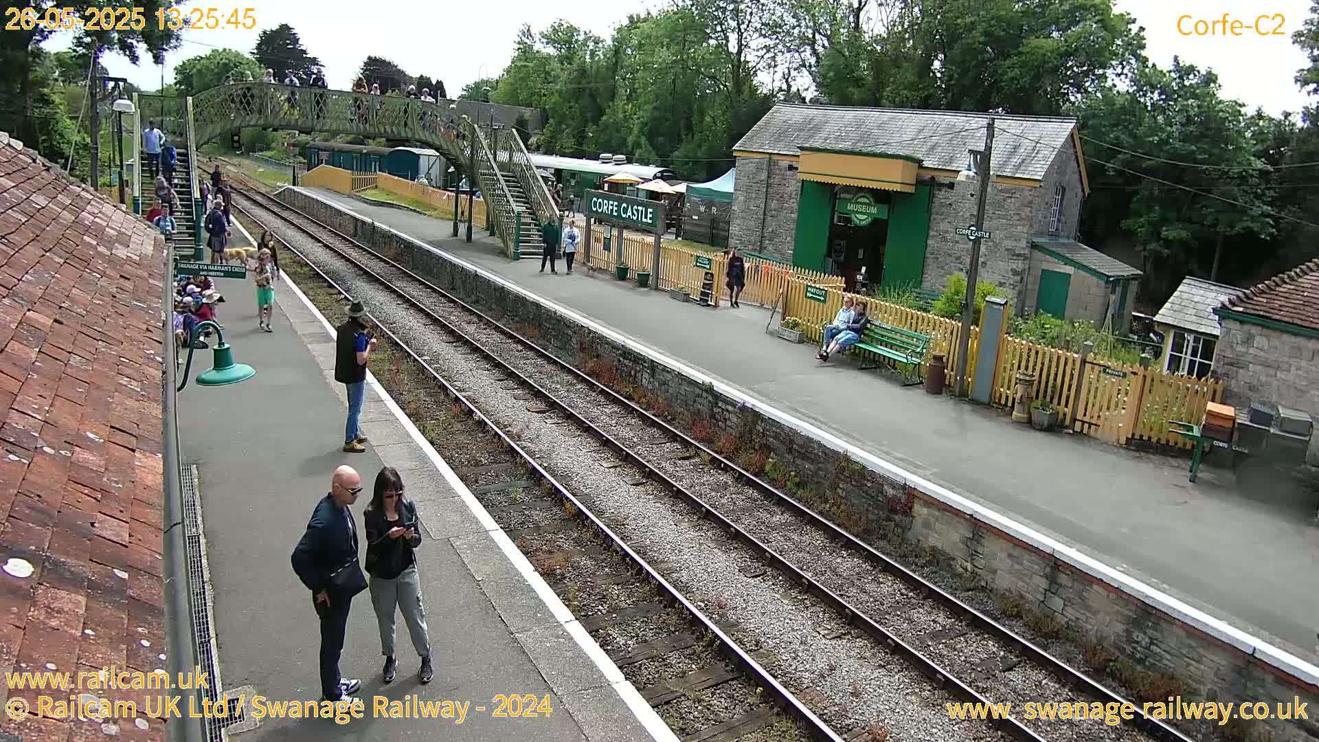 A sunny day at a train station with multiple people standing on the platform and a few sitting on benches, alongside railway tracks and a small building.