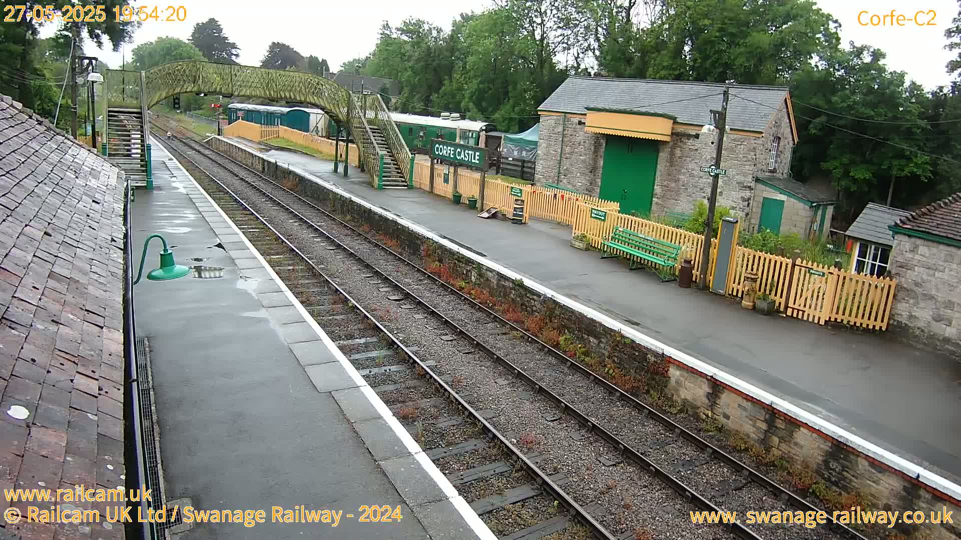 A wet train station platform with a railway track, a pedestrian bridge, and several stone buildings is visible on an overcast day.