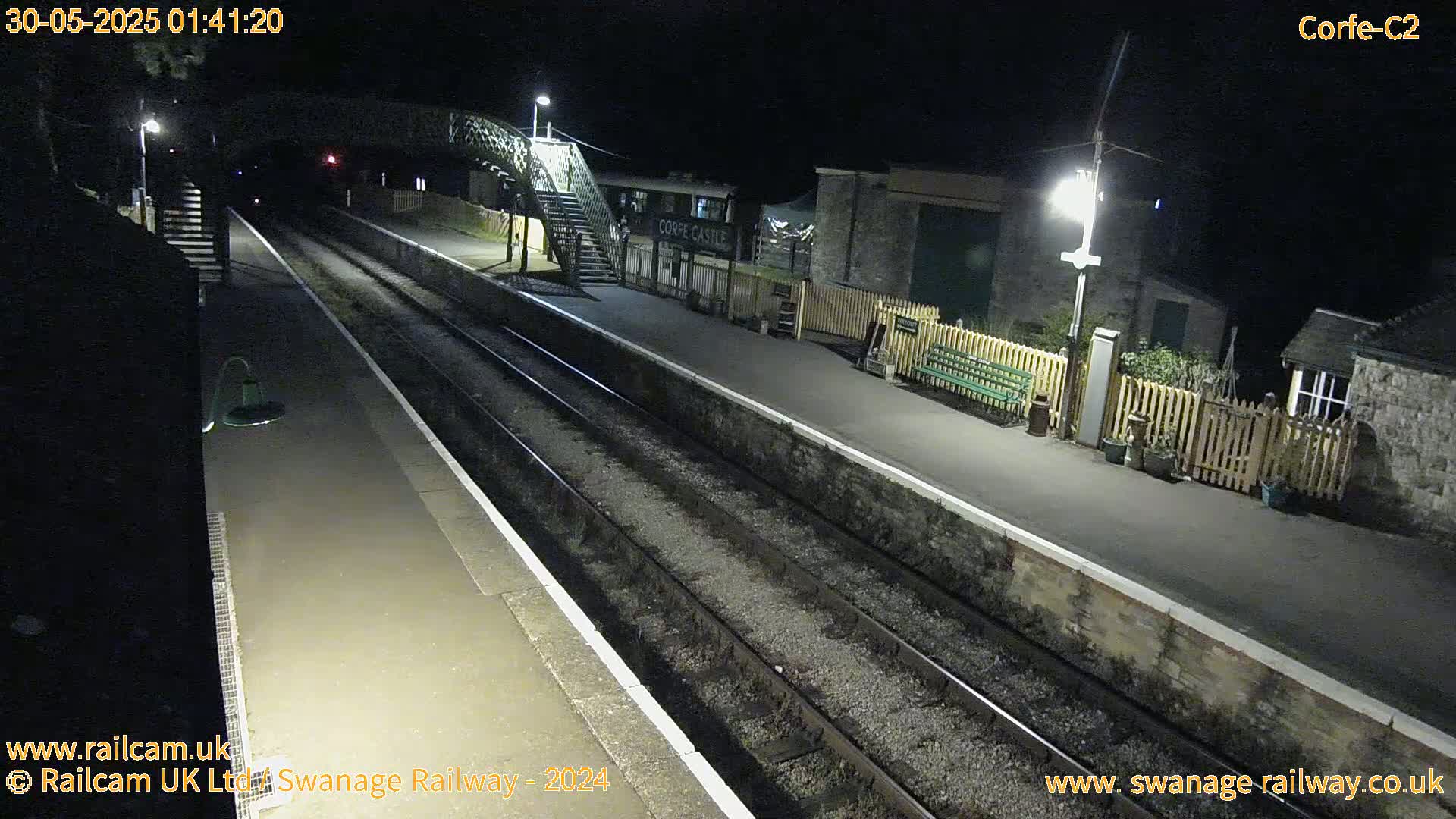 A nighttime view of a deserted train station platform with two sets of tracks, illuminated by several lamps.