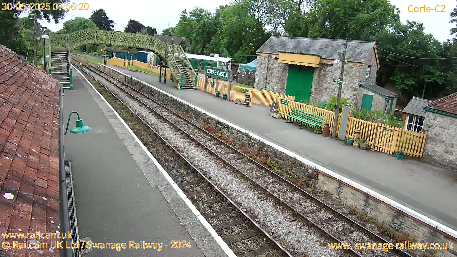 A train station platform with two railway tracks, a pedestrian footbridge, and stone buildings under an overcast sky.
