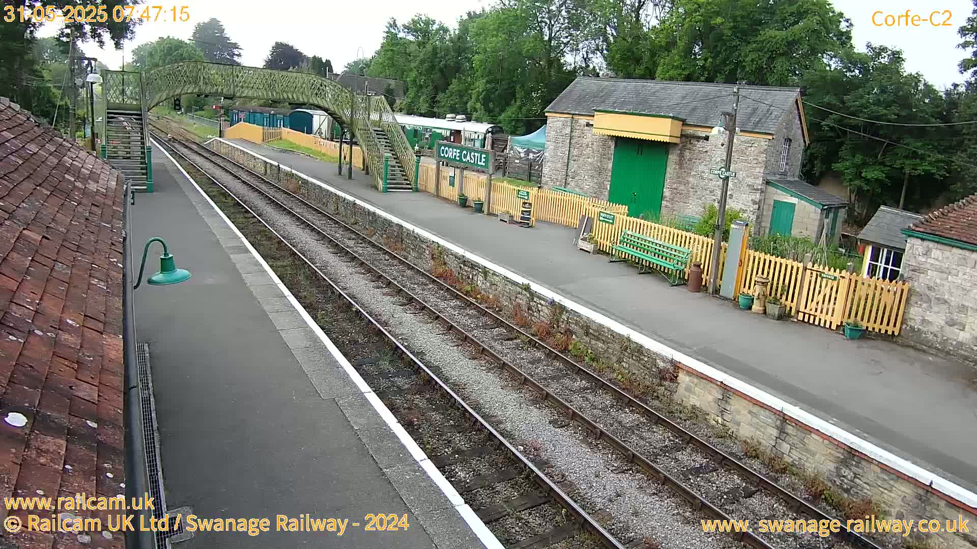 An overcast view of a train station platform with railway tracks, a pedestrian bridge, and stone buildings.