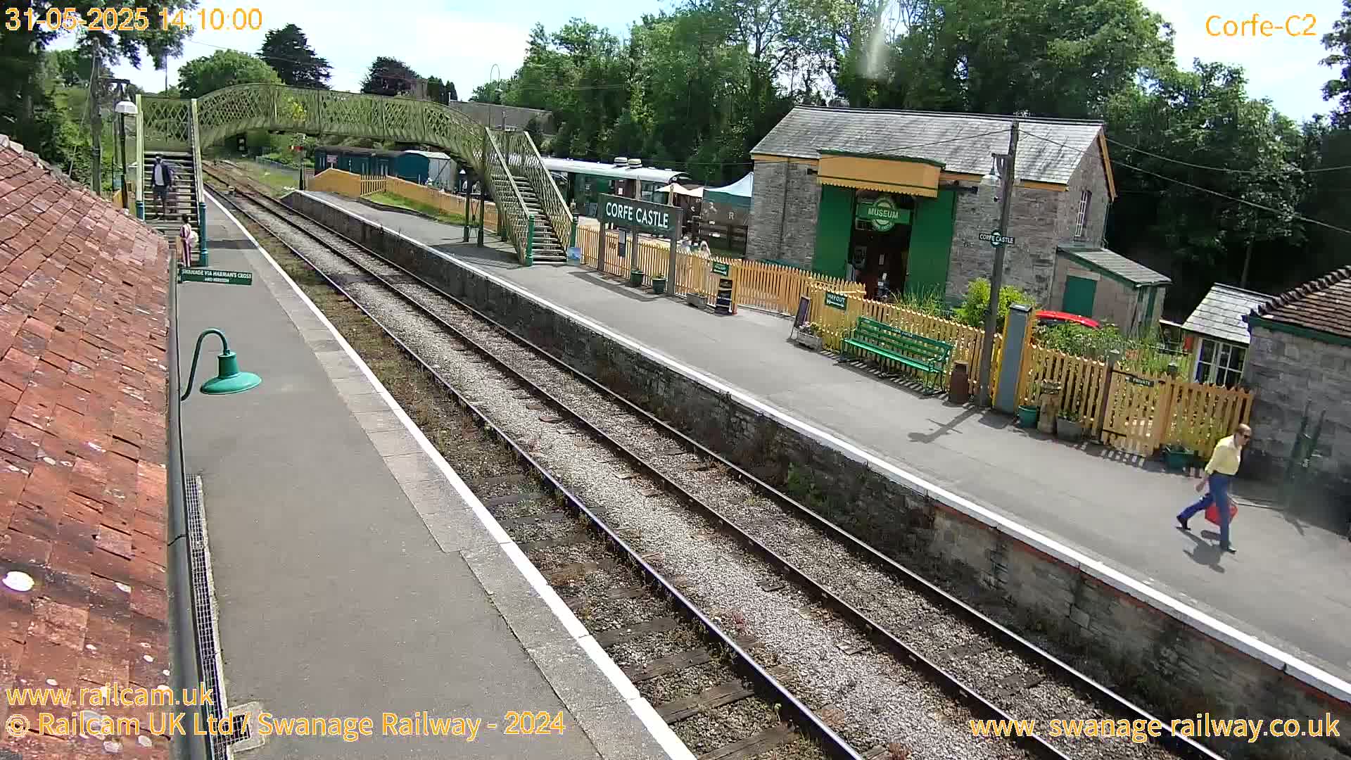 A sunny day at a train station shows a platform with two sets of tracks, a pedestrian bridge, and several buildings, including a museum.