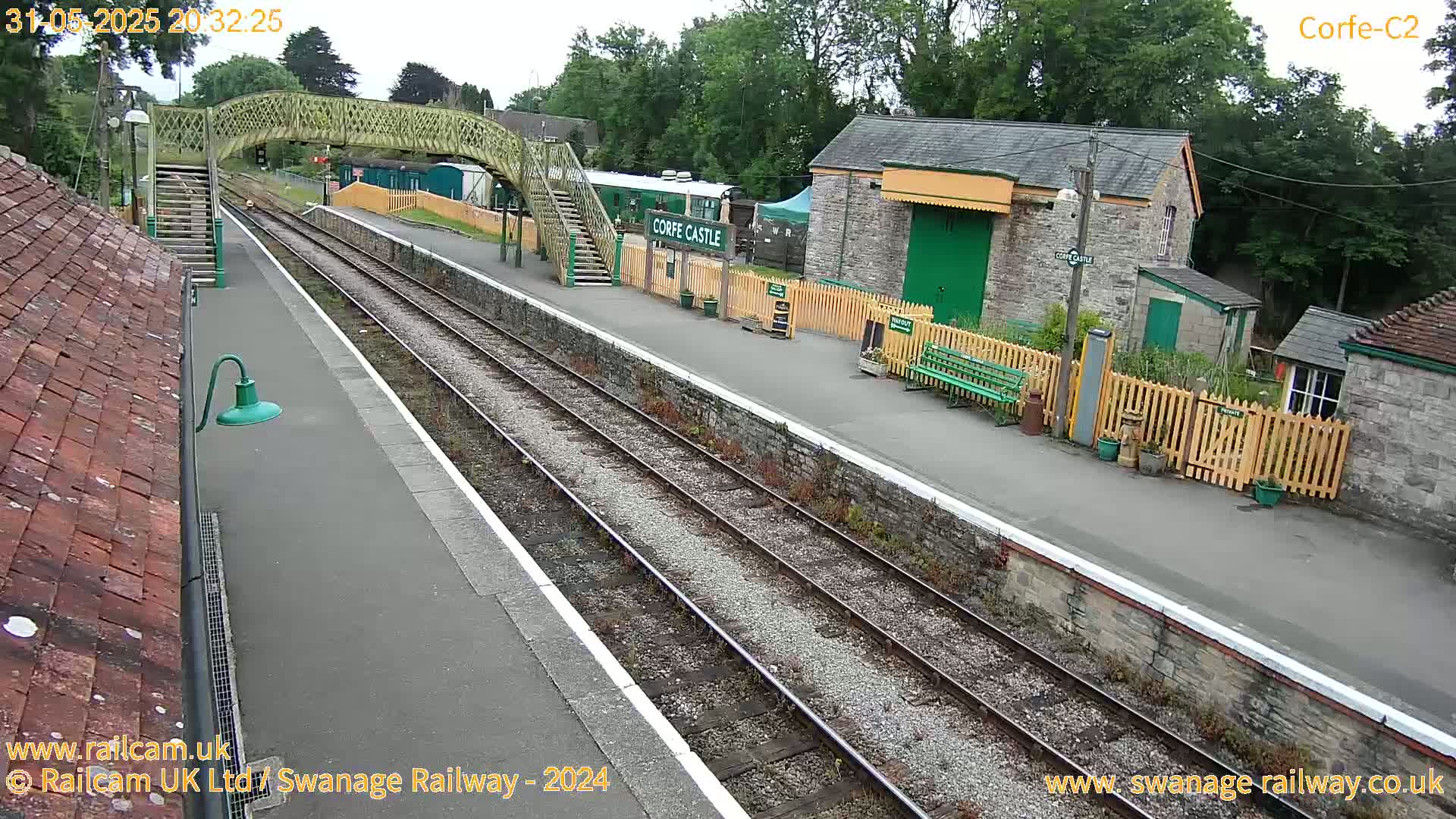 A railway station platform with train tracks, a stone building, a footbridge, and a small garden, under an overcast sky.