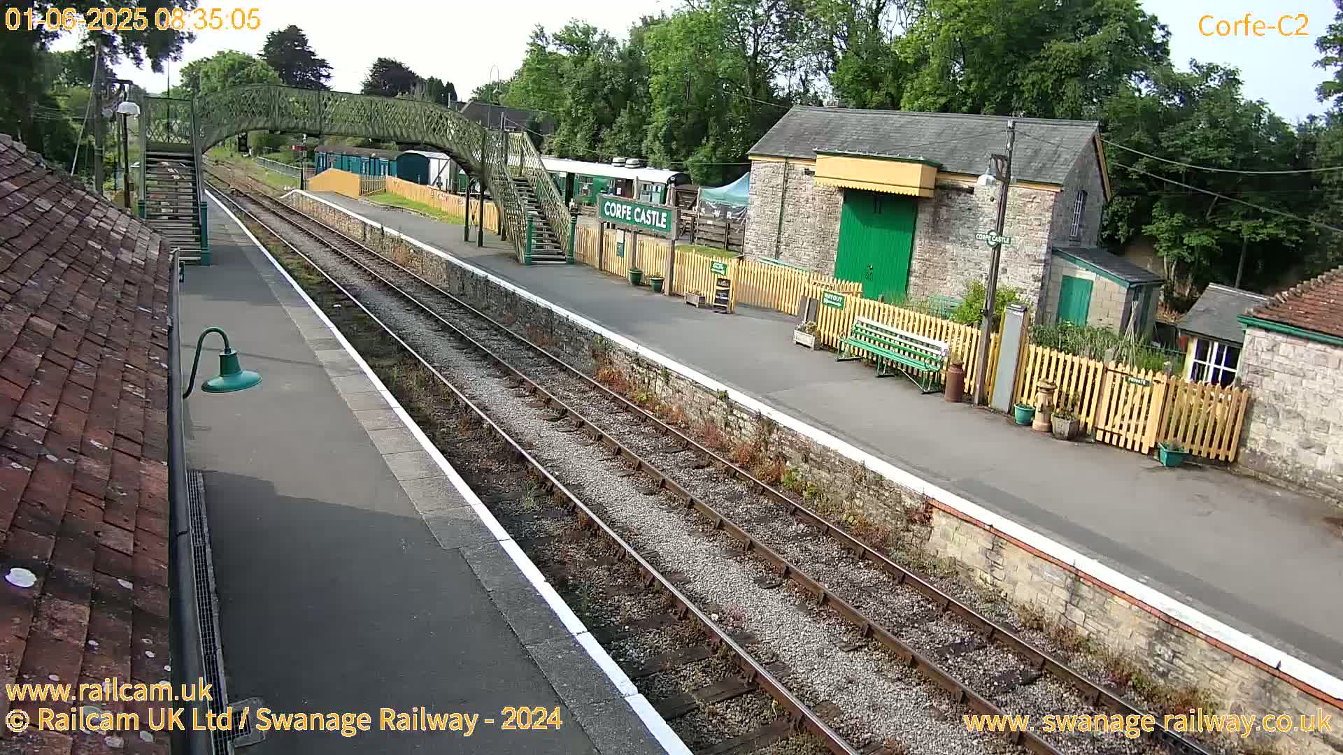 A train station platform with railway tracks, stone buildings, a footbridge, and a green-painted building under a sunny sky.