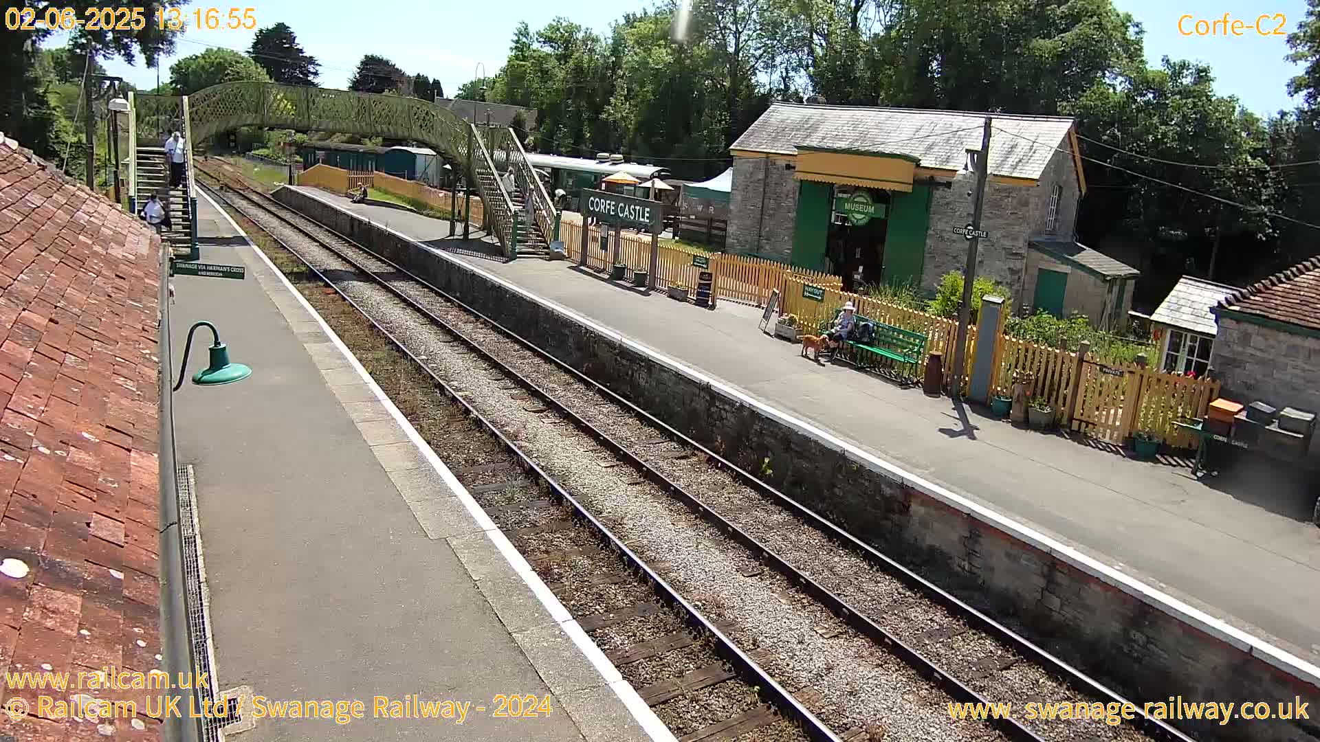 A sunny view of a train station platform with multiple tracks, a pedestrian bridge, and several buildings, including a small museum.