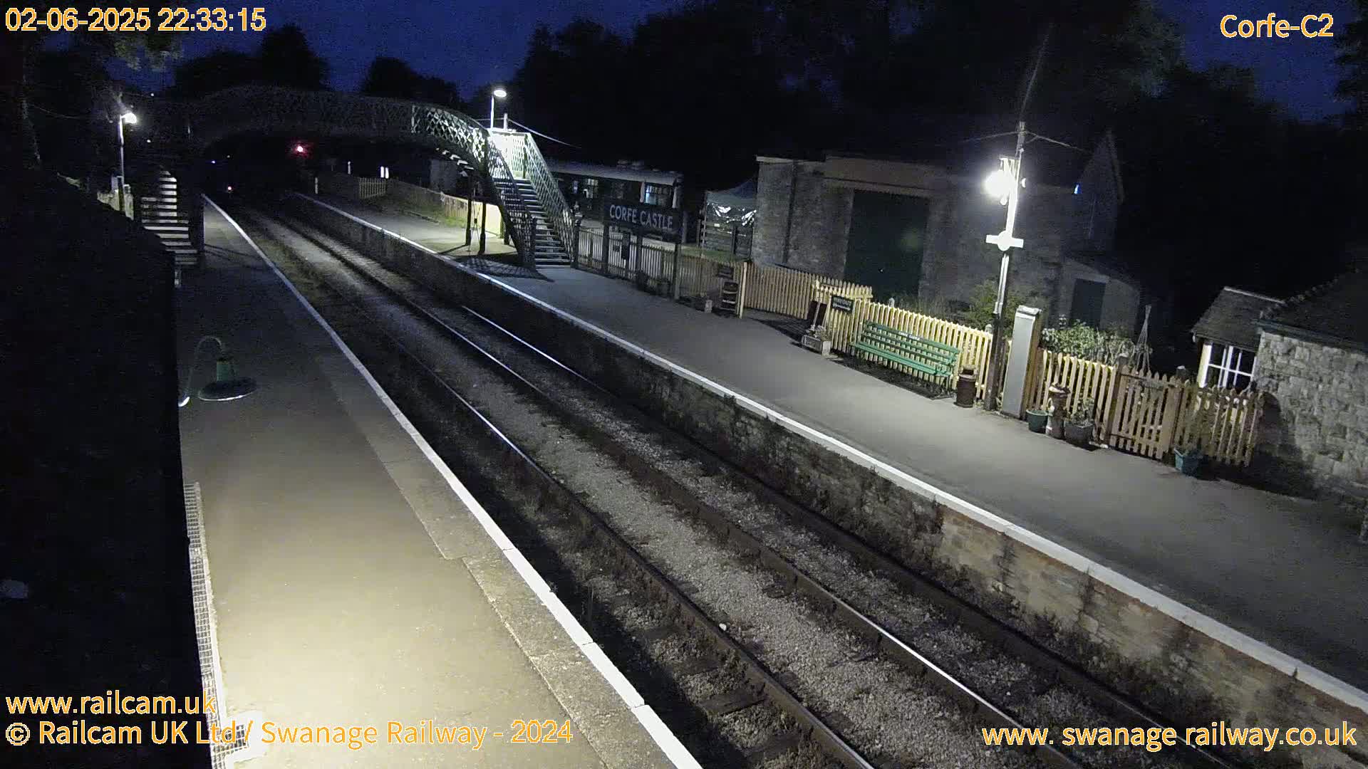 A nighttime view of a deserted train station platform with two sets of train tracks, a pedestrian bridge, and several buildings.