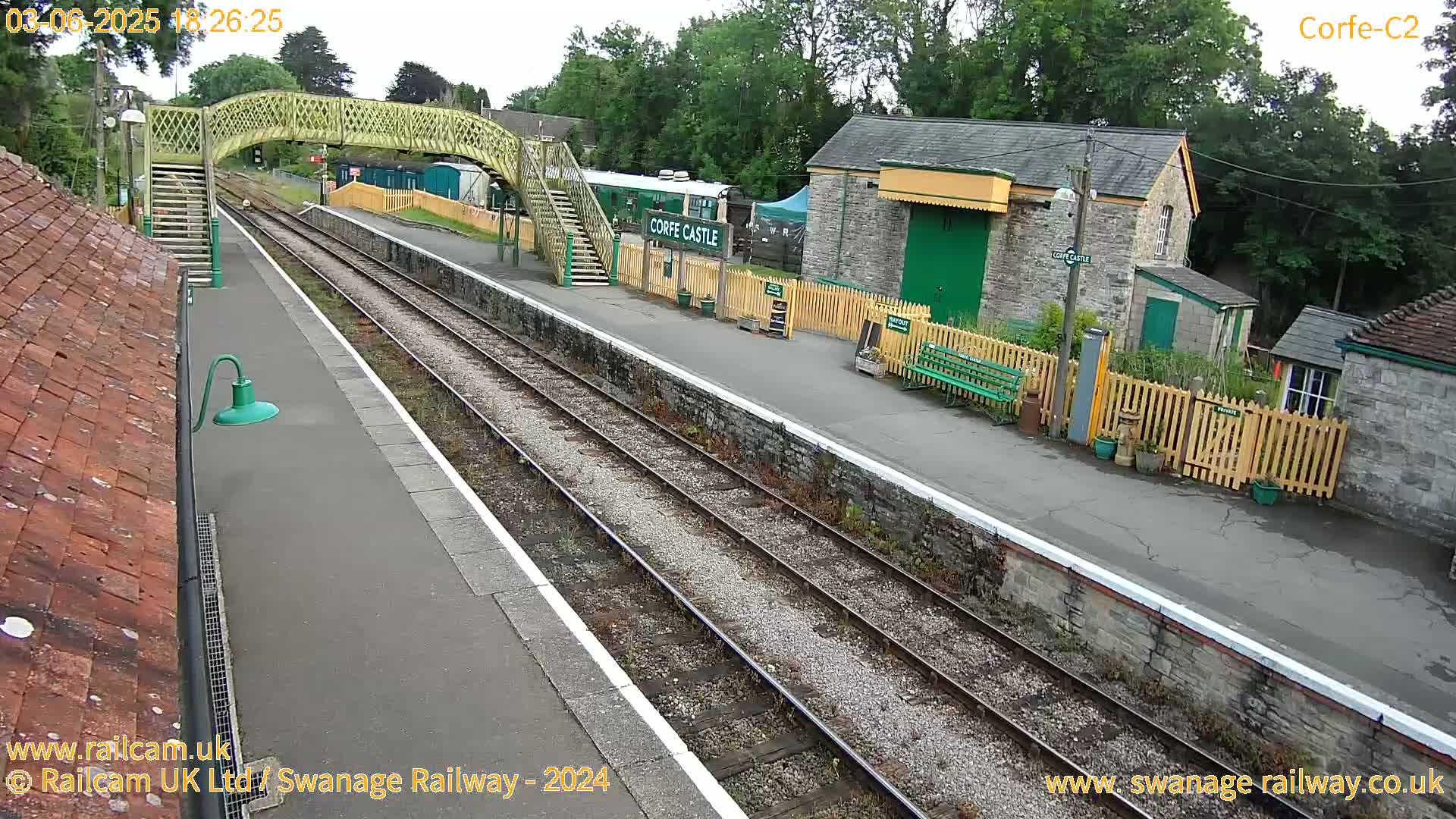 An overcast day at a train station shows a railway platform with tracks, a pedestrian bridge, and stone buildings.