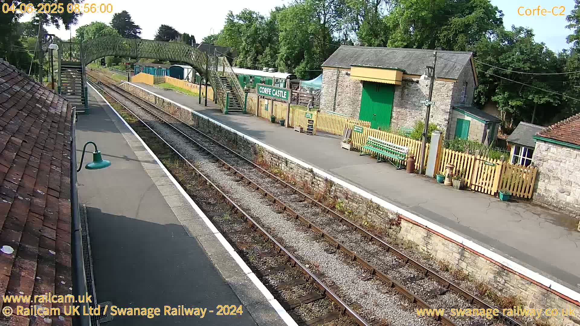 A train station platform with railway tracks, stone buildings, and a footbridge under a sunny sky.