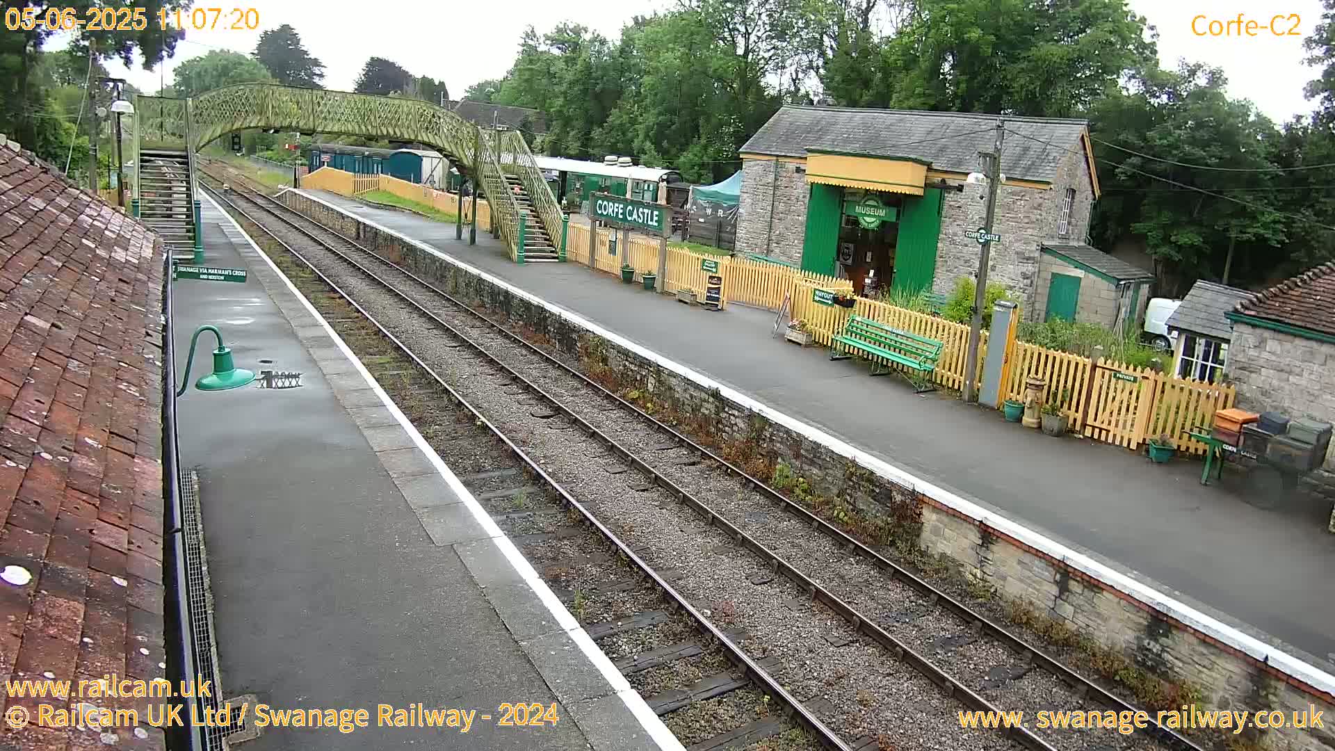 A railway station platform with train tracks, a pedestrian bridge, and a stone building under an overcast sky.