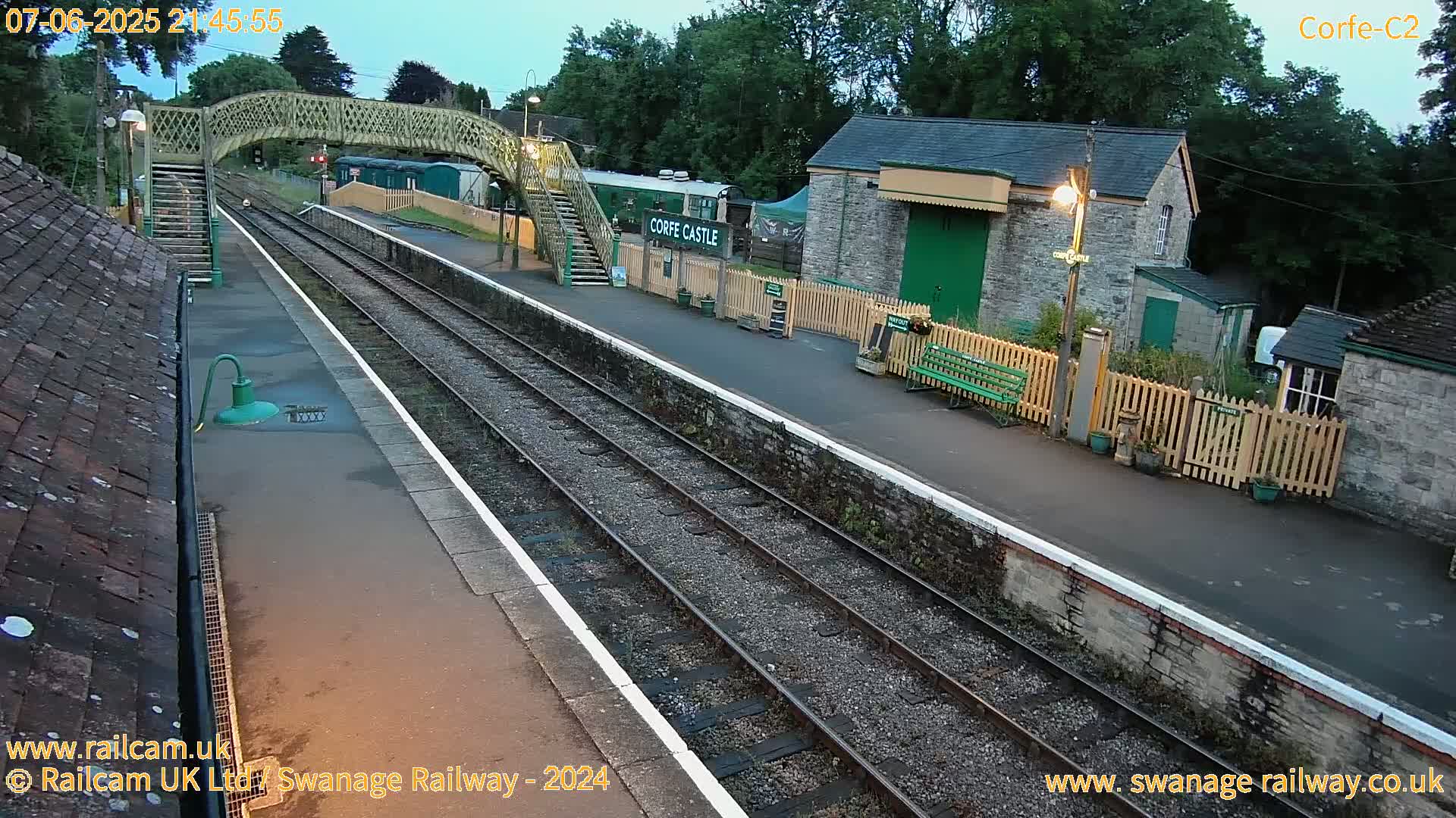 A train station platform at dusk, with a footbridge and a stone building visible under a mostly clear sky.