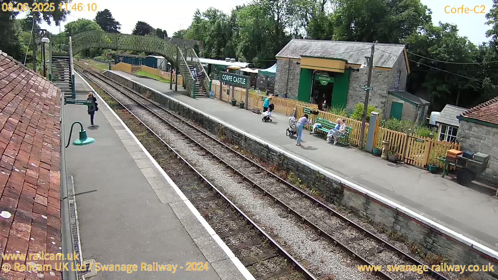 A sunny day at a train station with people walking and waiting near stone buildings and tracks.