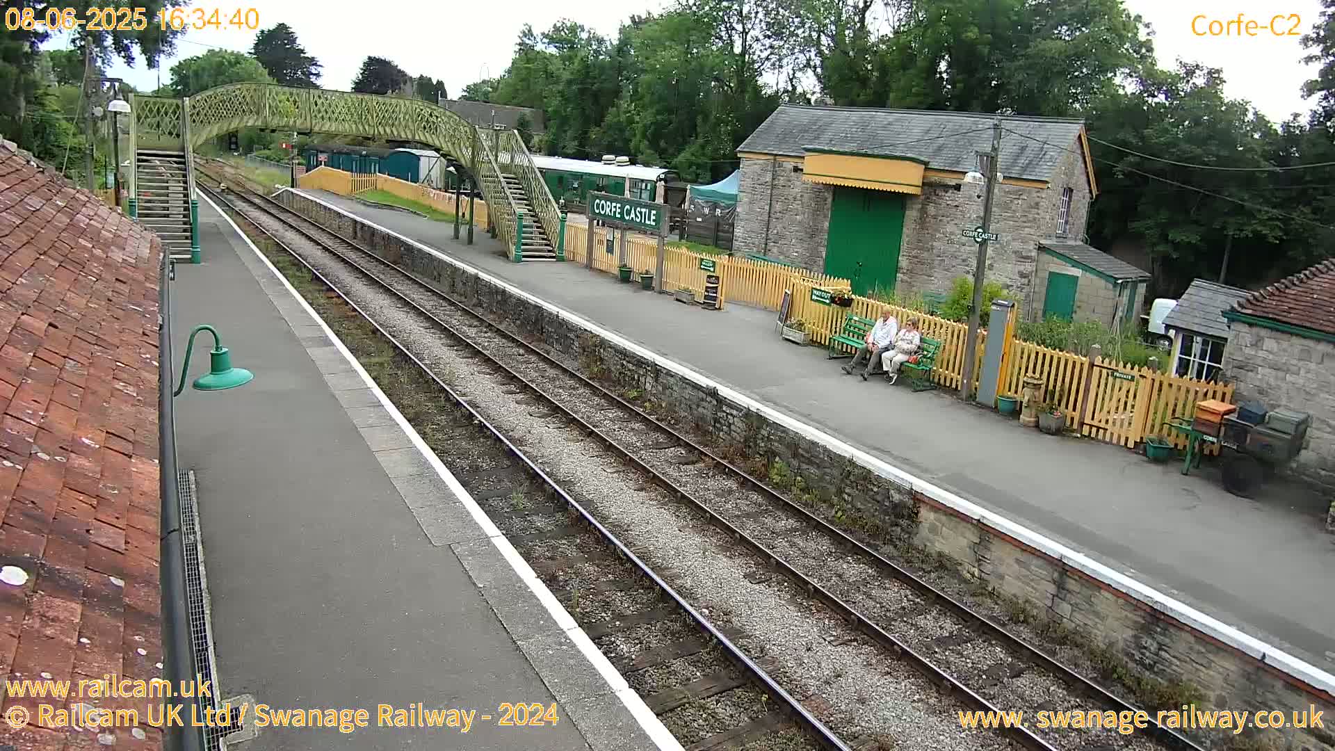 A train station platform with a footbridge, stone buildings, and two people sitting on a bench, under an overcast sky.