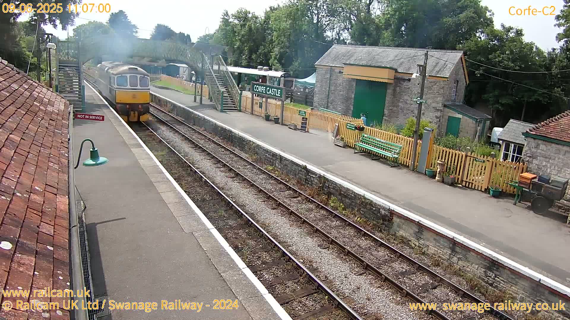 A train is departing a station on a sunny day, passing stone buildings and a wooden fence.