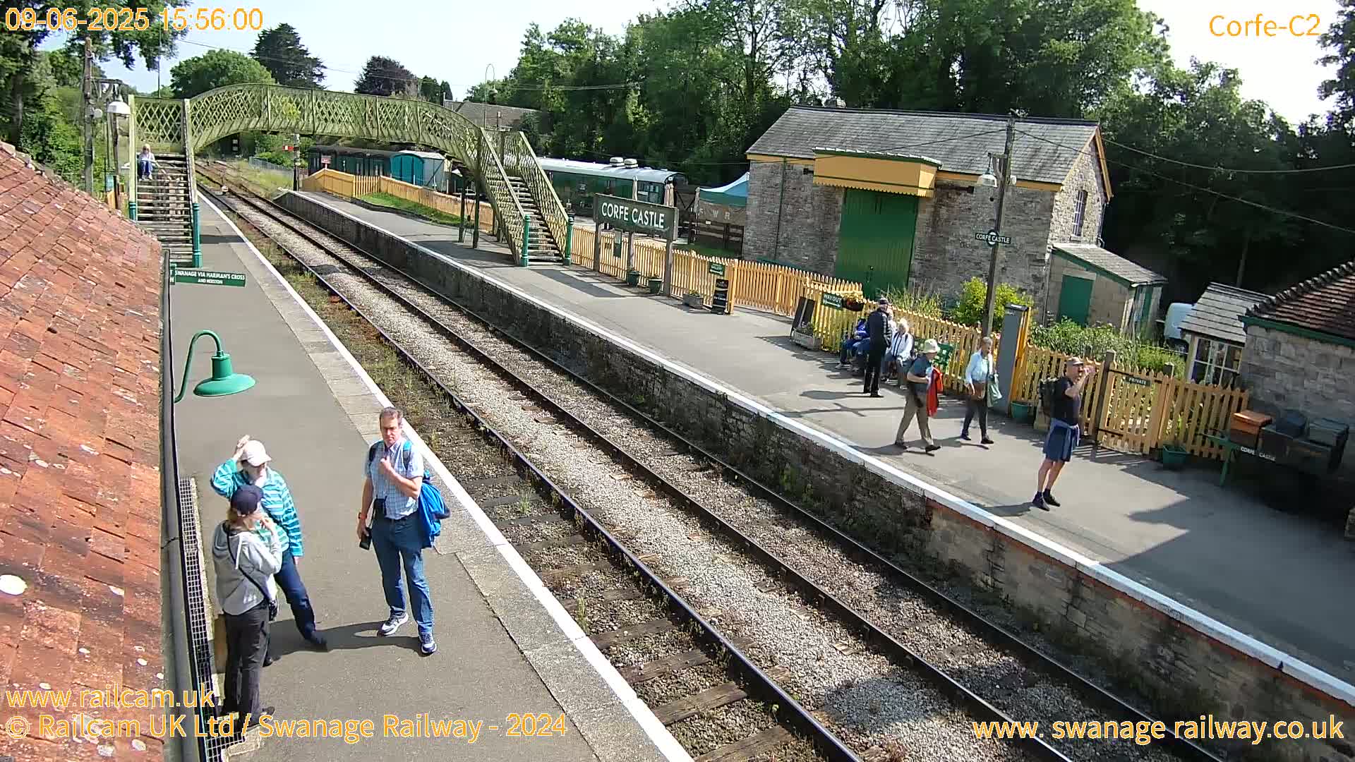 A sunny day at a train station shows a few people standing on the platform,  a train partially visible in the background, and several buildings nearby.