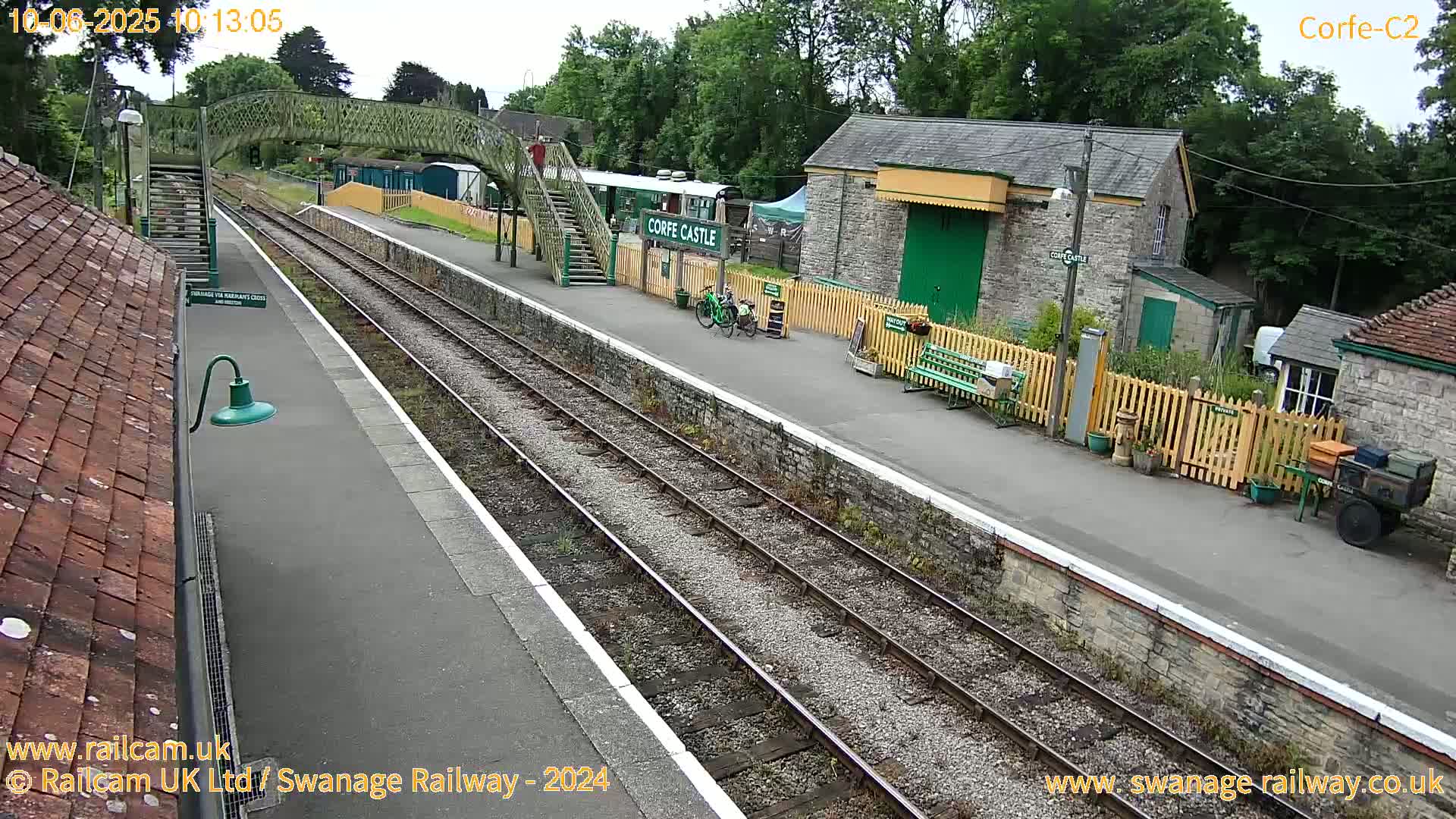 A train station platform with railway tracks, stone buildings, a footbridge, and some bicycles under an overcast sky.