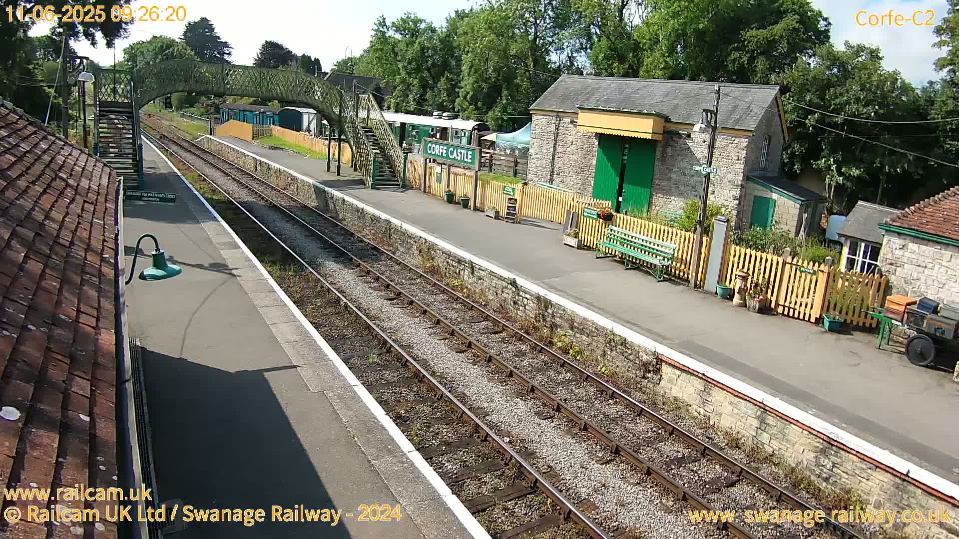 A sunny day at a train station with a stone building, a footbridge, and railway tracks.