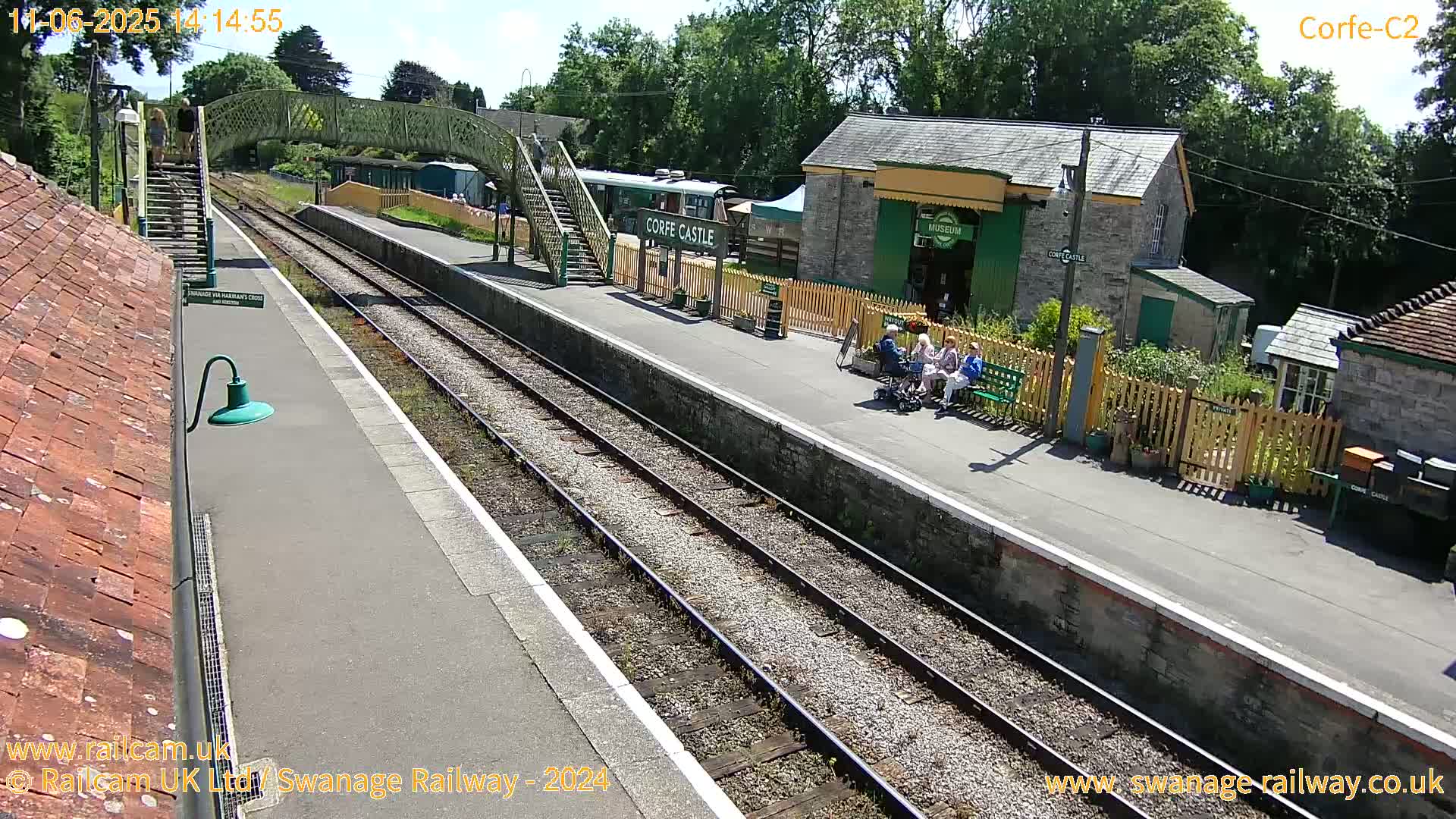 A sunny view of a train station platform with several people sitting on benches, a train car visible, and various buildings including a museum.