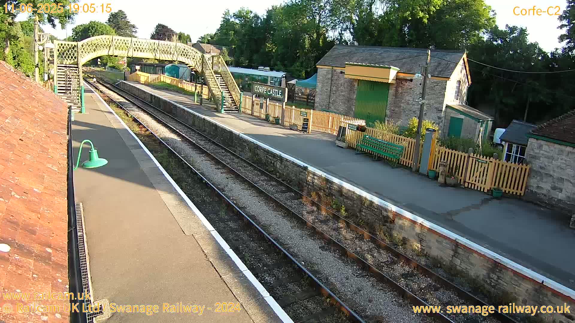 A train station with a pedestrian bridge, stone buildings, and train tracks under sunny conditions.