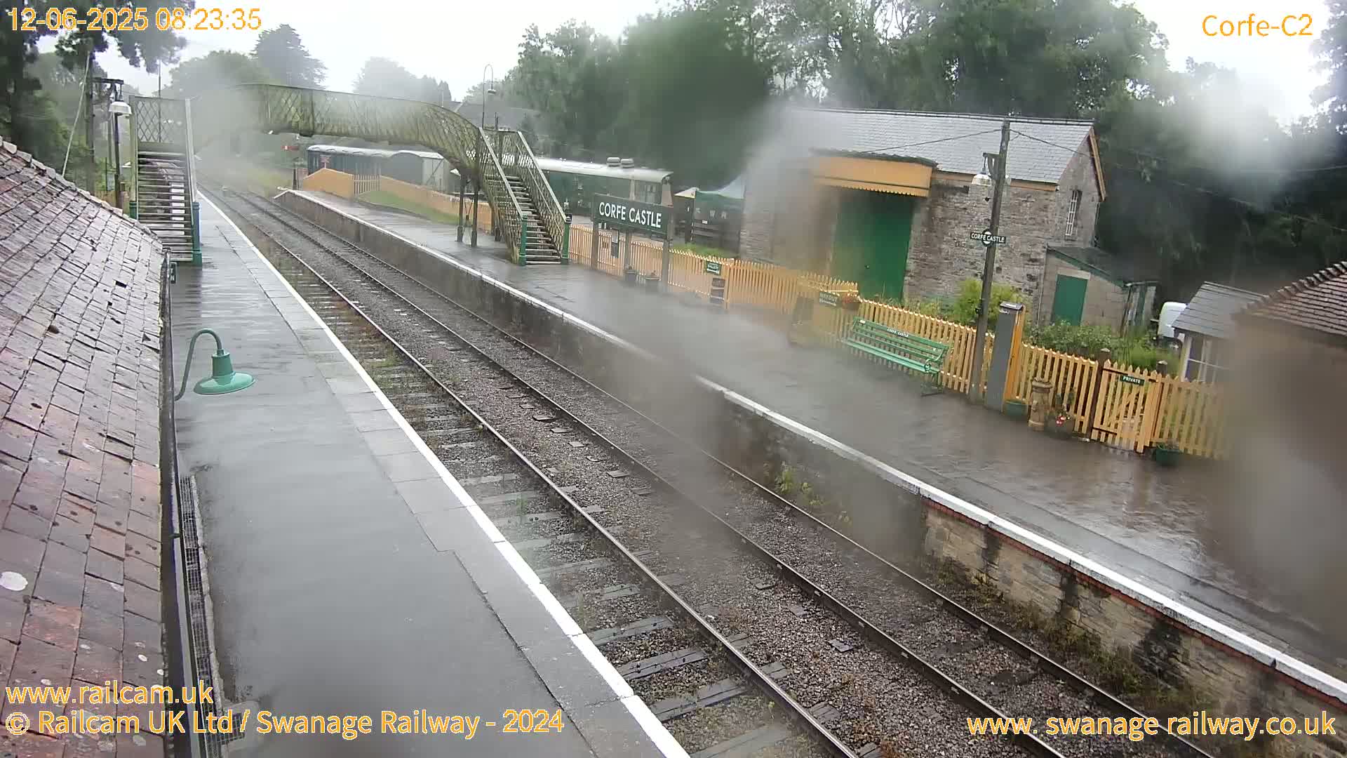 A rain-soaked railway station platform with tracks, a footbridge, and station buildings is seen under a gray sky.