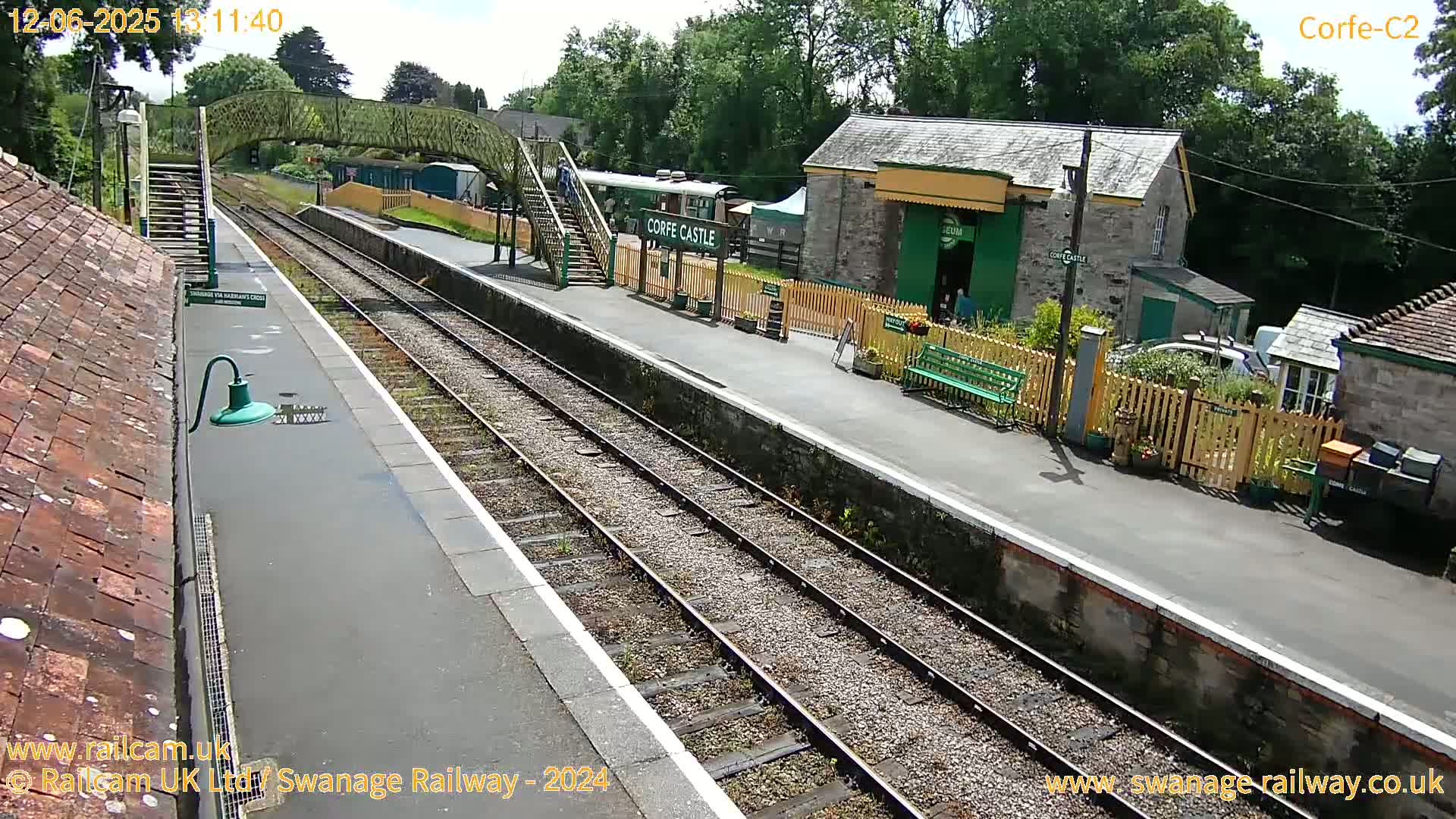 A train station platform with a train visible, several buildings, and a footbridge on a sunny day.